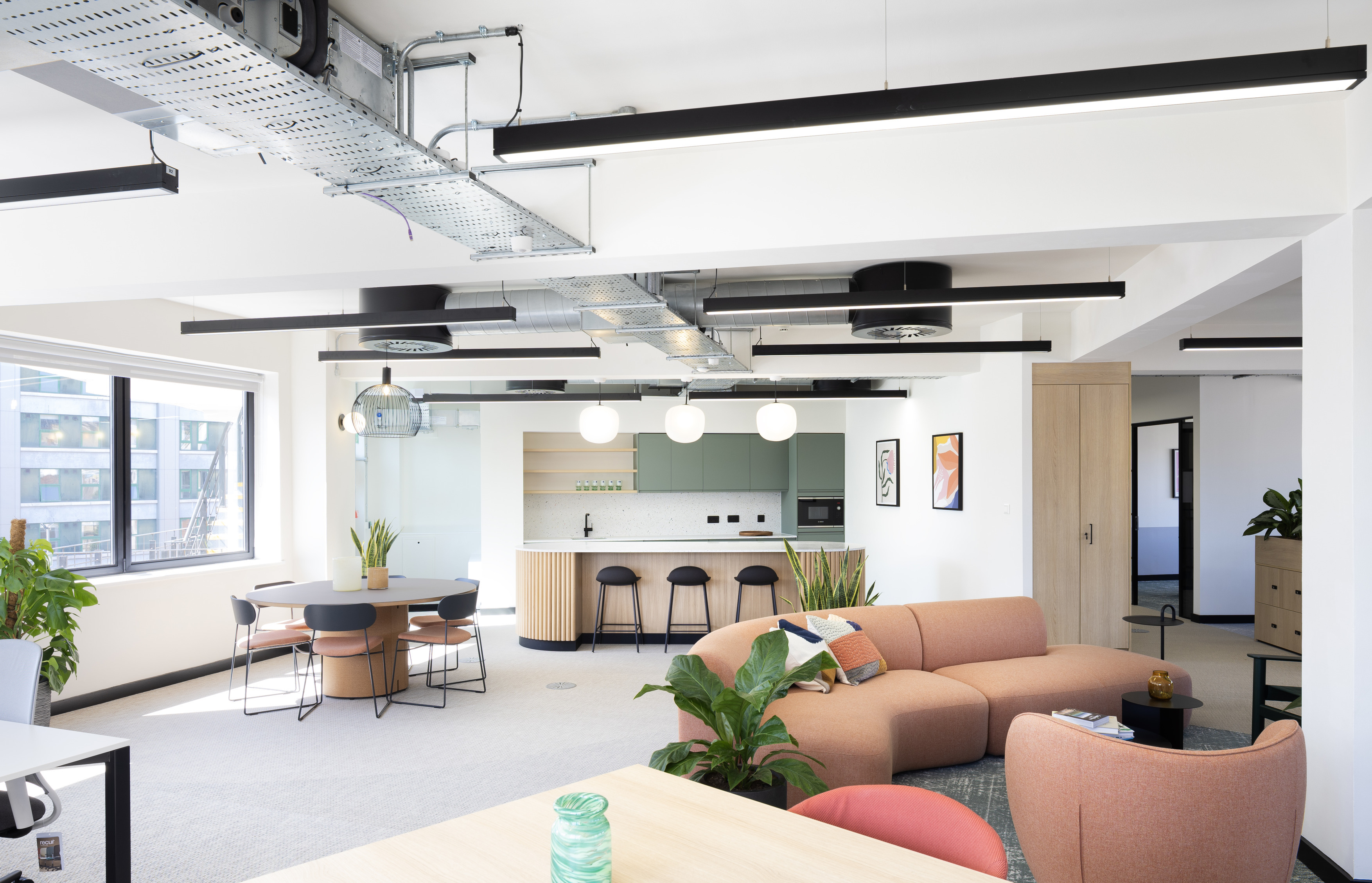 A modern open-plan office featuring industrial-style exposed ceiling ducts and sleek black linear light fixtures. The space includes a kitchen area with light wood cabinetry, white countertops and black bar stools. A round dining table with four chairs sits near a large window that floods the room with natural light. The foreground showcases a peach-coloured L-shaped sofa surrounded by green plants and small tables, creating a relaxed lounge area. Framed artwork adds visual interest to the neutral-toned walls.