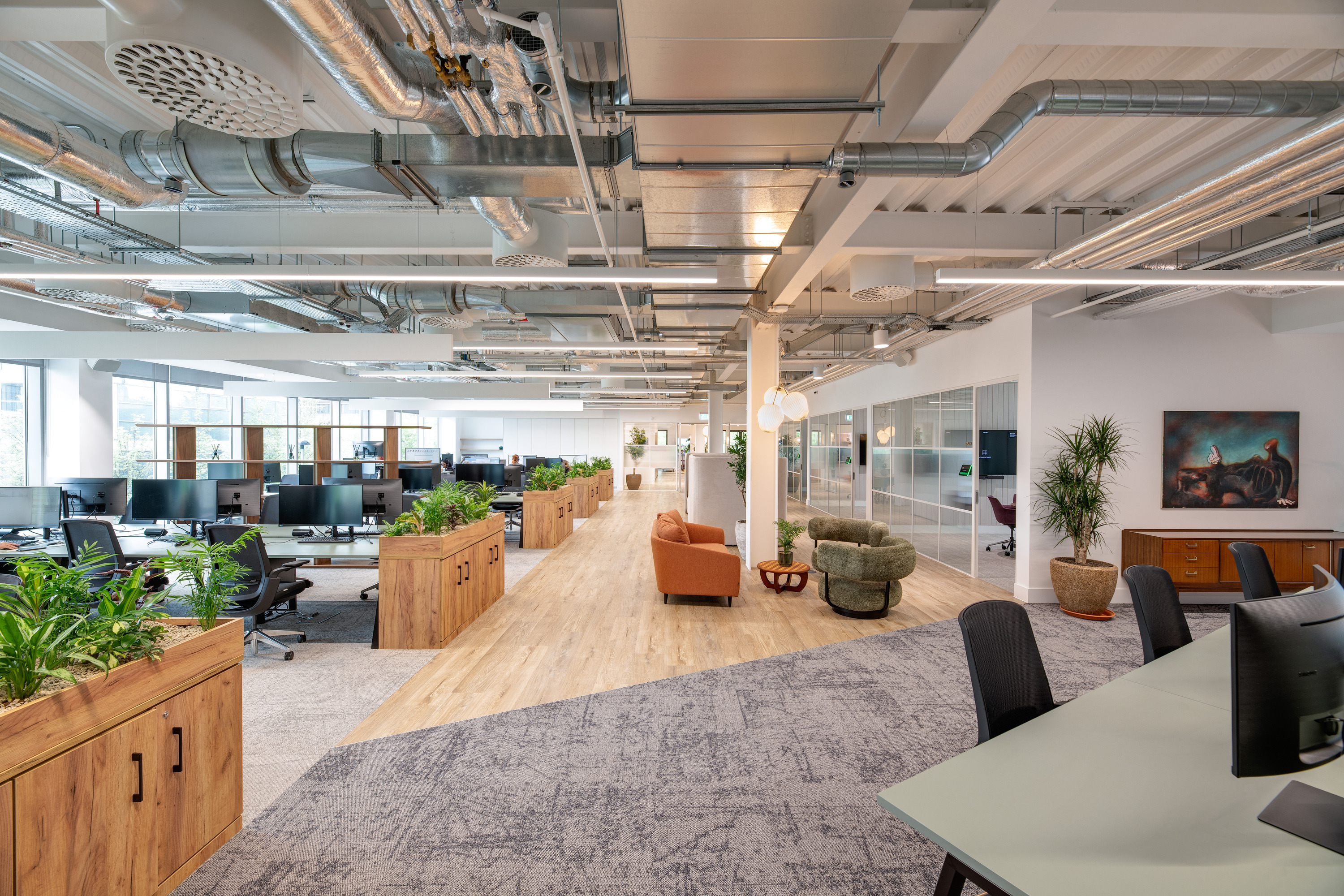 Bright, open plan office interior with an exposed ceiling showing silver ductwork and services, white beams and linear lighting. Rows of desks with multiple monitors and task chairs sit to the left, separated by long wooden planters filled with green plants. A central walkway with light wood flooring runs through the space, leading to a soft seating area with upholstered armchairs and a small table. On the right, a glass partition encloses meeting rooms with visible chairs inside, alongside a sideboard, potted plants and wall art. The space combines neutral tones, natural materials and greenery, with carpeted zones around the workstations and a clean, contemporary finish throughout.