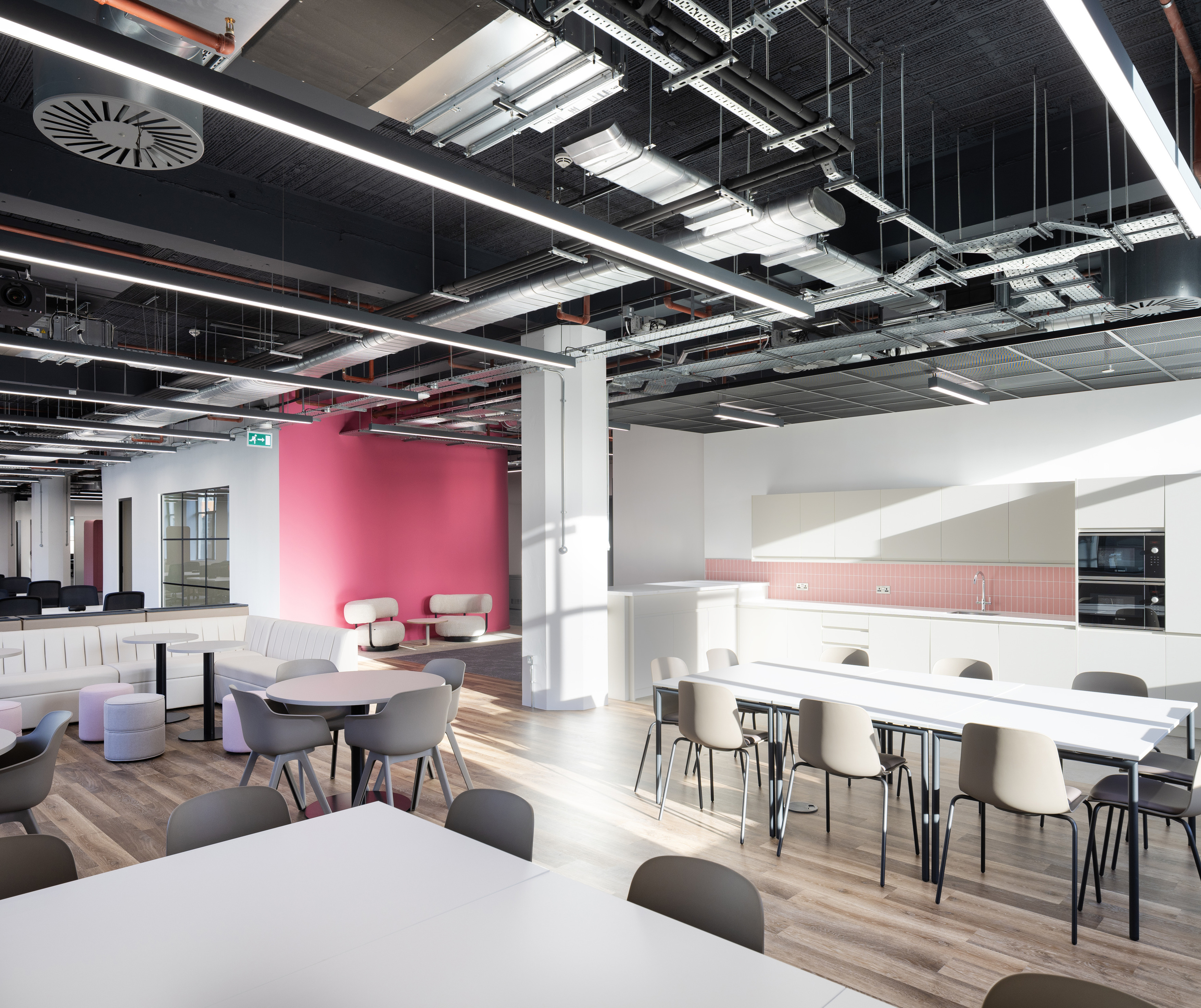 Modern open plan office breakout and kitchen area with a clean, contemporary fit out. The space features long white dining tables with light grey chairs in the foreground, alongside smaller round tables and soft seating to the left. At the back right, a white kitchenette includes integrated appliances, cabinets and a pink tiled splashback. A bold pink feature wall sits behind two white lounge chairs, adding contrast to the otherwise neutral palette. The ceiling is exposed, showing ventilation, cable trays and linear LED lighting, while large windows and overhead lights create a bright, evenly lit environment. The flooring is a mix of wood effect vinyl and darker carpet in the seating area.