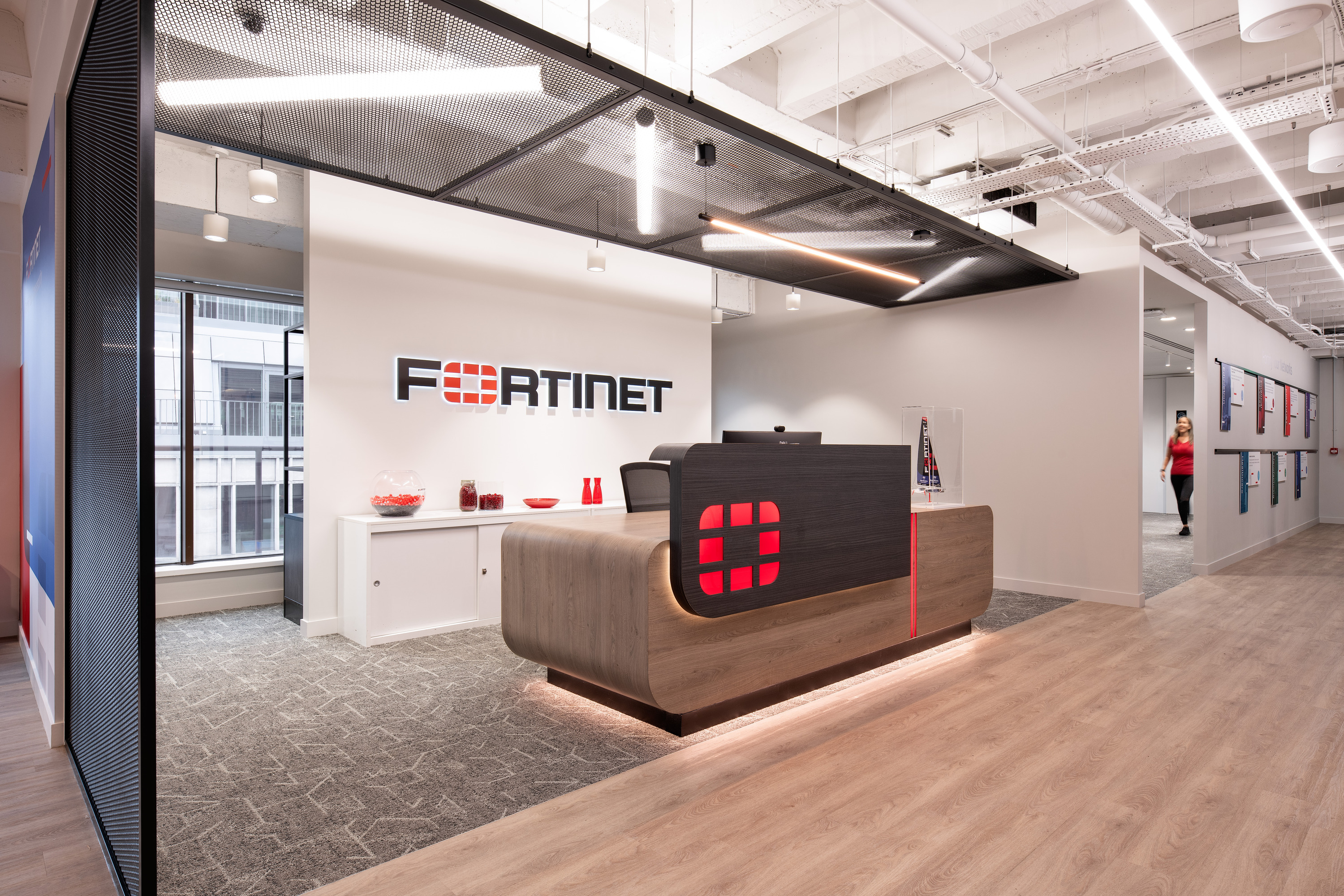 Modern office reception area featuring a curved timber and black-fronted reception desk with illuminated red Fortinet branding. The Fortinet logo is displayed on a white feature wall behind the desk, with large windows to the left providing natural light. Exposed ceilings, suspended mesh panels and linear LED lighting create an industrial-style finish, while a corridor with framed displays extends to the right. A person is visible in the background walking through the office.