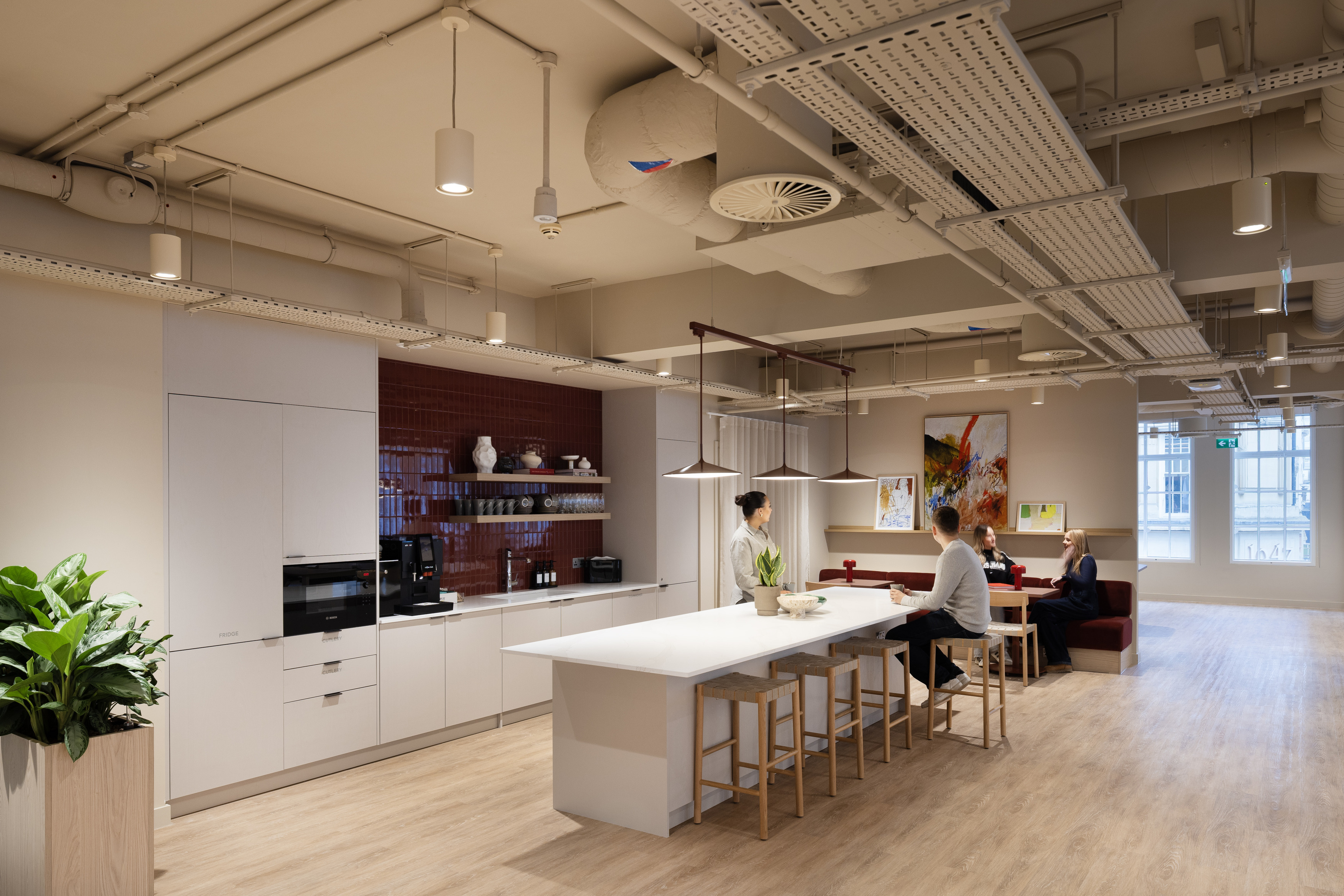 Modern open plan office with a bright, minimalist design featuring a central communal table with high stools, integrated coffee station and natural wood flooring. Exposed ceiling services and suspended lighting create an industrial aesthetic, complemented by large windows for natural light and greenery for a biophilic touch. Flexible workstations are visible in the background, promoting collaboration and contemporary workplace design