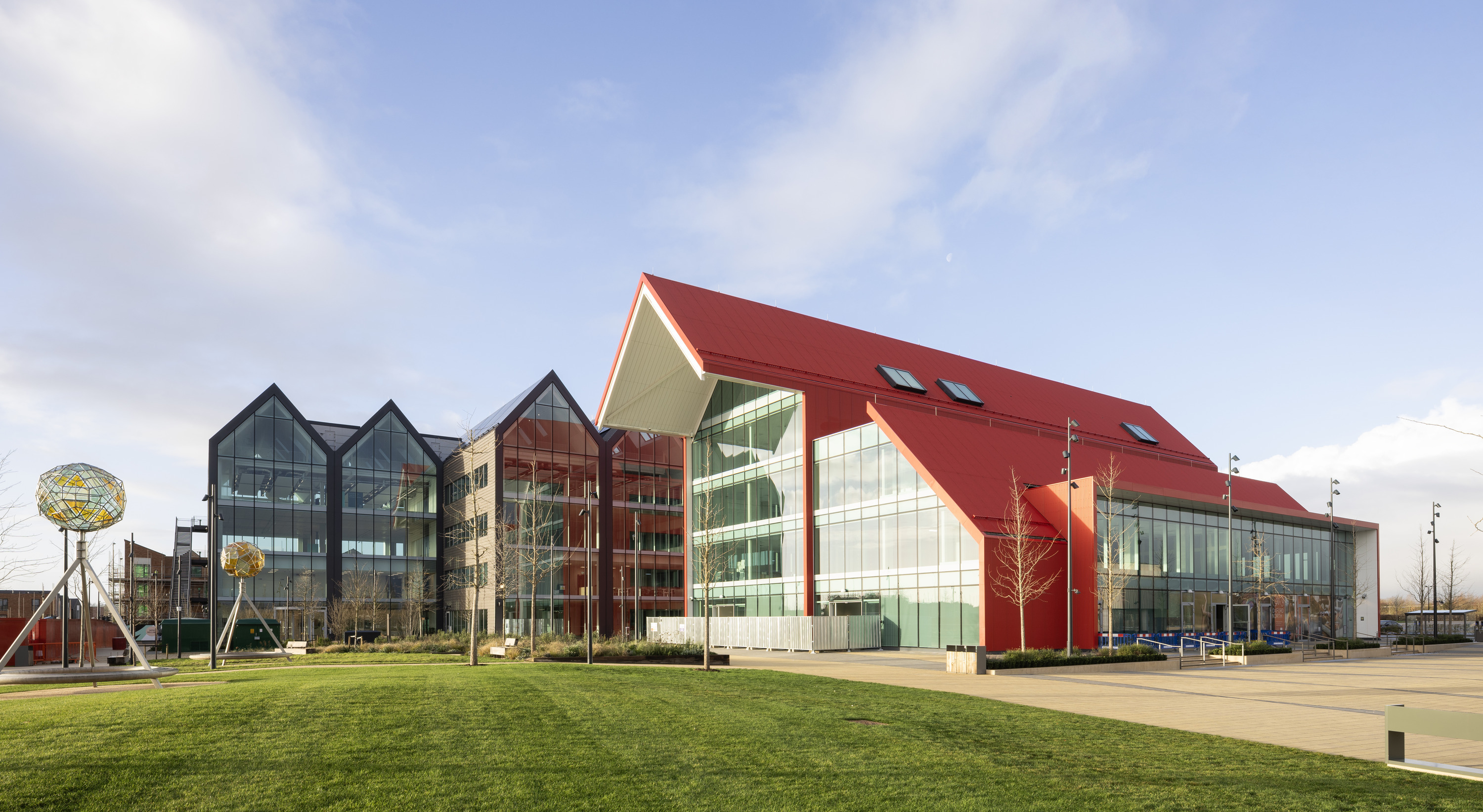 Modern campus-style building with a red pitched roof and large glass façades, connected to a darker grey, multi-gabled glass structure on the left. The buildings sit beside a wide paved walkway and a neatly maintained green lawn in the foreground. Sculptural metal frames holding spherical artworks stand on the grass. The scene is brightly lit by sunlight under a clear blue sky with light clouds, highlighting the clean lines, reflective windows and contemporary architectural design.