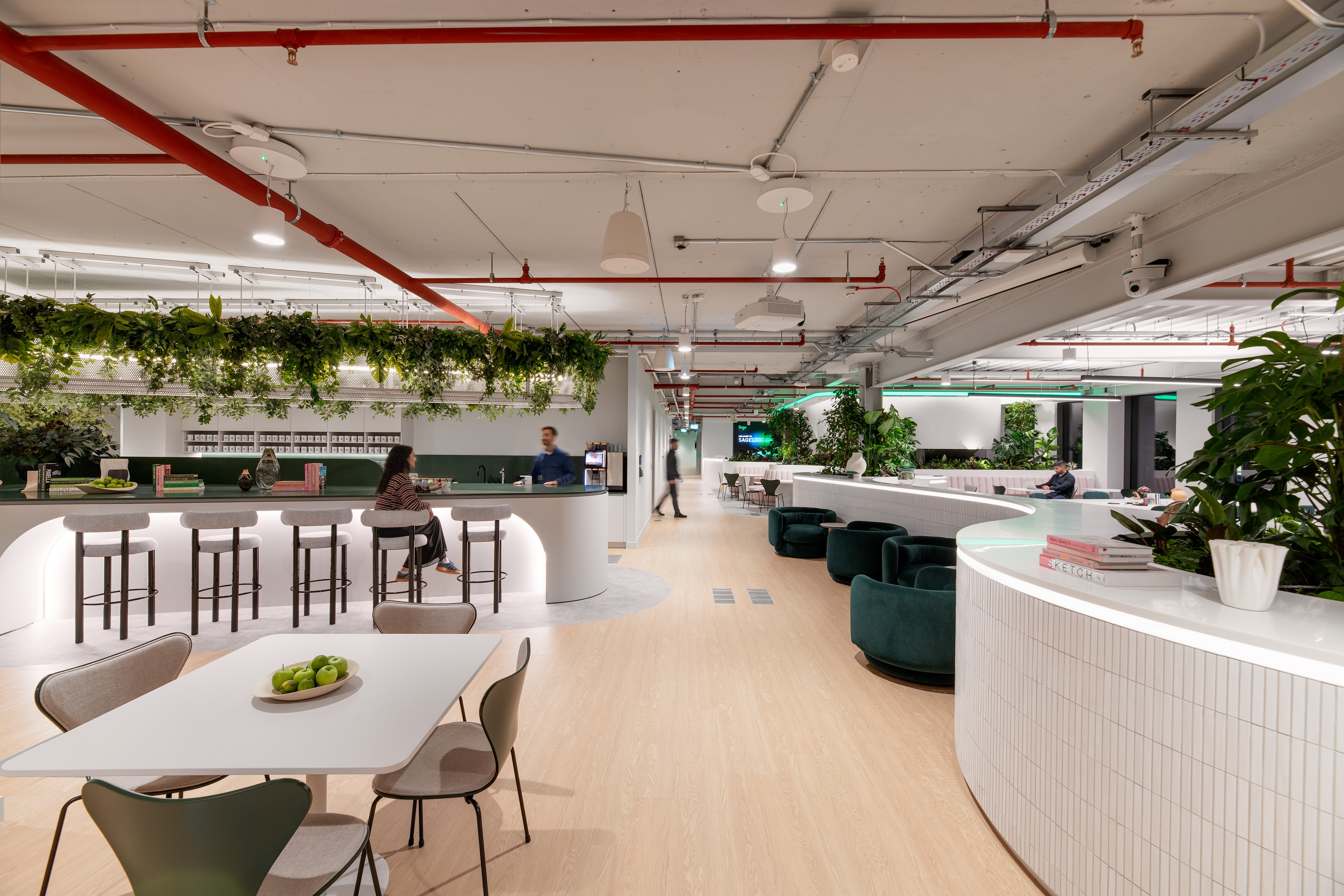 A spacious modern workplace with light wood flooring, white curved counters and abundant greenery. A central bar-style area with stools sits beneath a ceiling feature covered in hanging plants. People are seated at tables and lounge chairs throughout the open space. Overhead pipes and lighting are exposed, and the setting has a bright, contemporary feel.