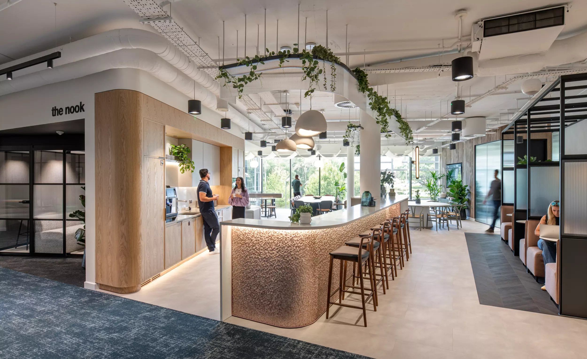 A modern, open-concept office space anchored by a central bar-style counter surrounded by stools. The ceiling showcases exposed ductwork and hanging plants, contributing to the contemporary aesthetic. To the left, a glass-partitioned area labeled the nook offers extra seating and workspace. Large windows in the background fill the room with natural light, while people move through the space, adding a sense of energy and flow.