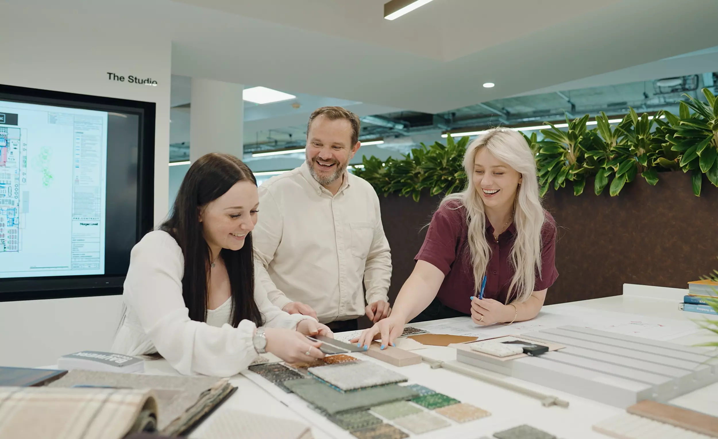 Three individuals are gathered around a table covered with material samples including fabric swatches and tiles. They are engaged in a collaborative discussion. Behind them, a large screen displays a layout titled The Studio. A shelf with green plants adds a touch of natural decor to the setting