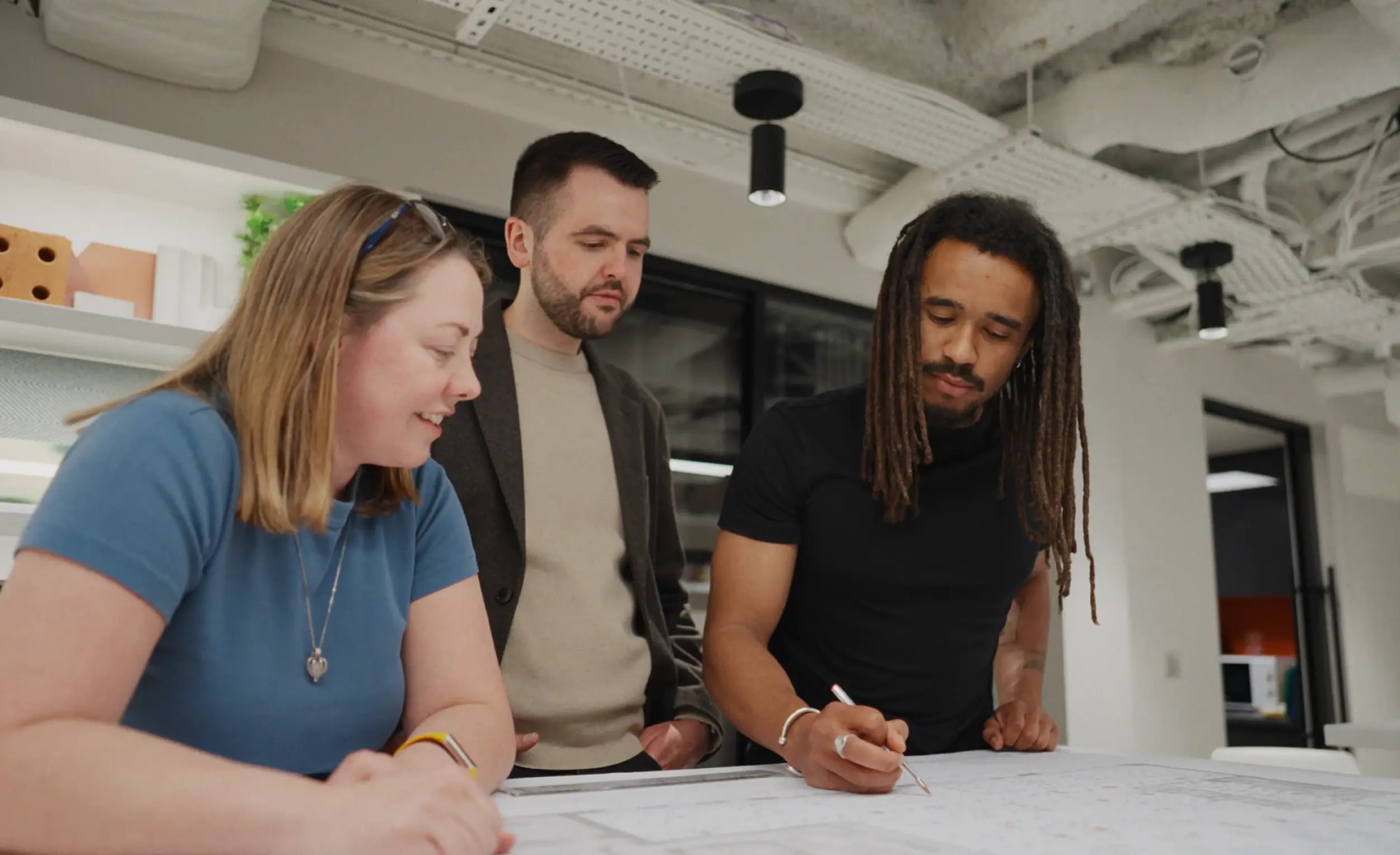 Three people gathered around a table reviewing large printed plans in a modern office. One person is writing on the documents while the other two look on. The space features exposed services and cable trays on the ceiling, black pendant lights, white shelving with sample materials and a glass partition in the background. The image focuses on collaboration and design review within a contemporary workplace setting.  Provide your feedback on BizChat