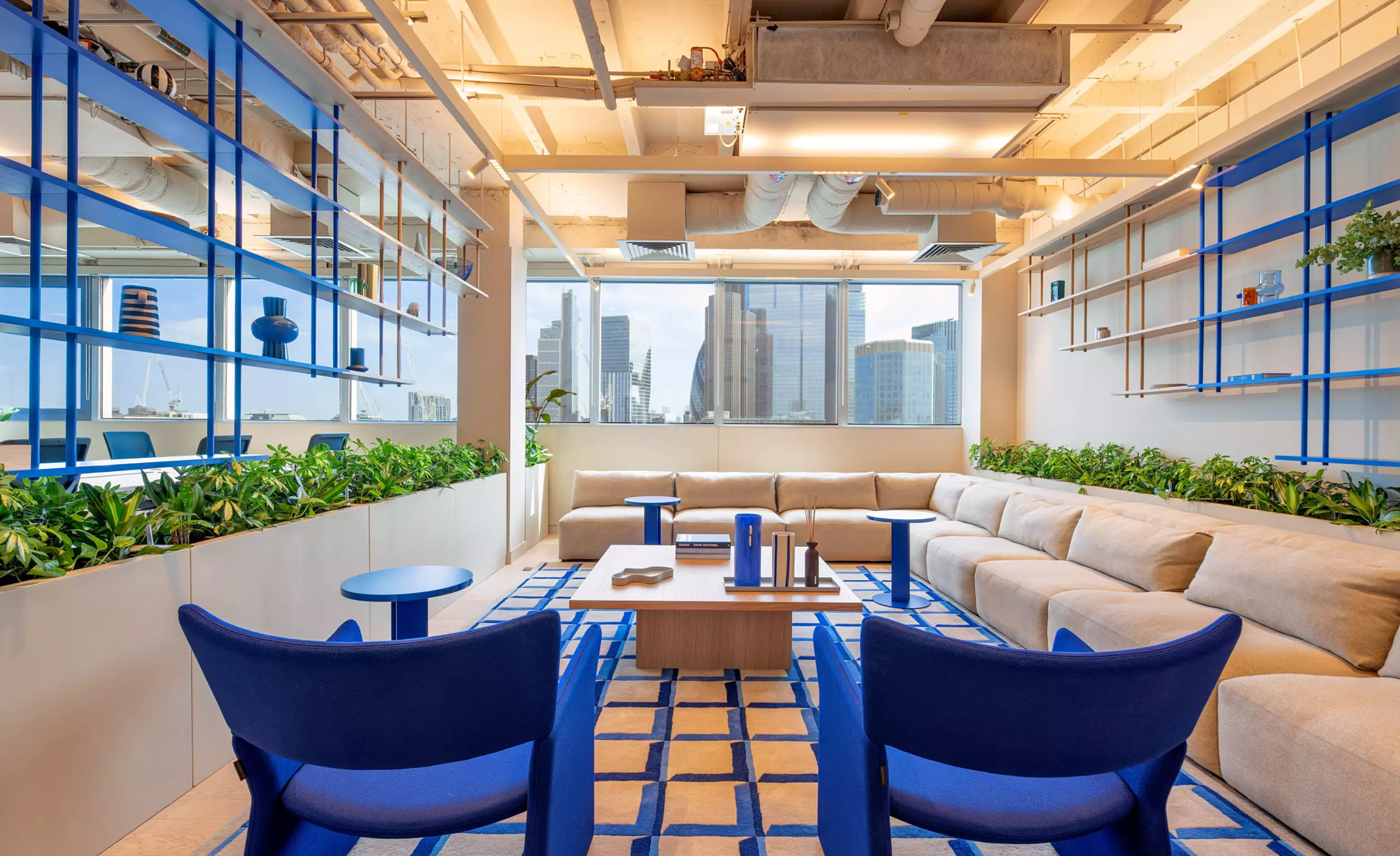 A contemporary office lounge with an open, airy feel. The space is defined by large windows that frame a city skyline, flooding the room with natural light. Beige sectional sofas form an inviting L-shape, complemented by two blue armchairs and several small tables for casual meetings or relaxation. Blue shelving units line the walls, displaying decorative items that add personality to the space. Green plants soften the industrial look created by exposed ceiling pipes and ducts, blending comfort with modern design.