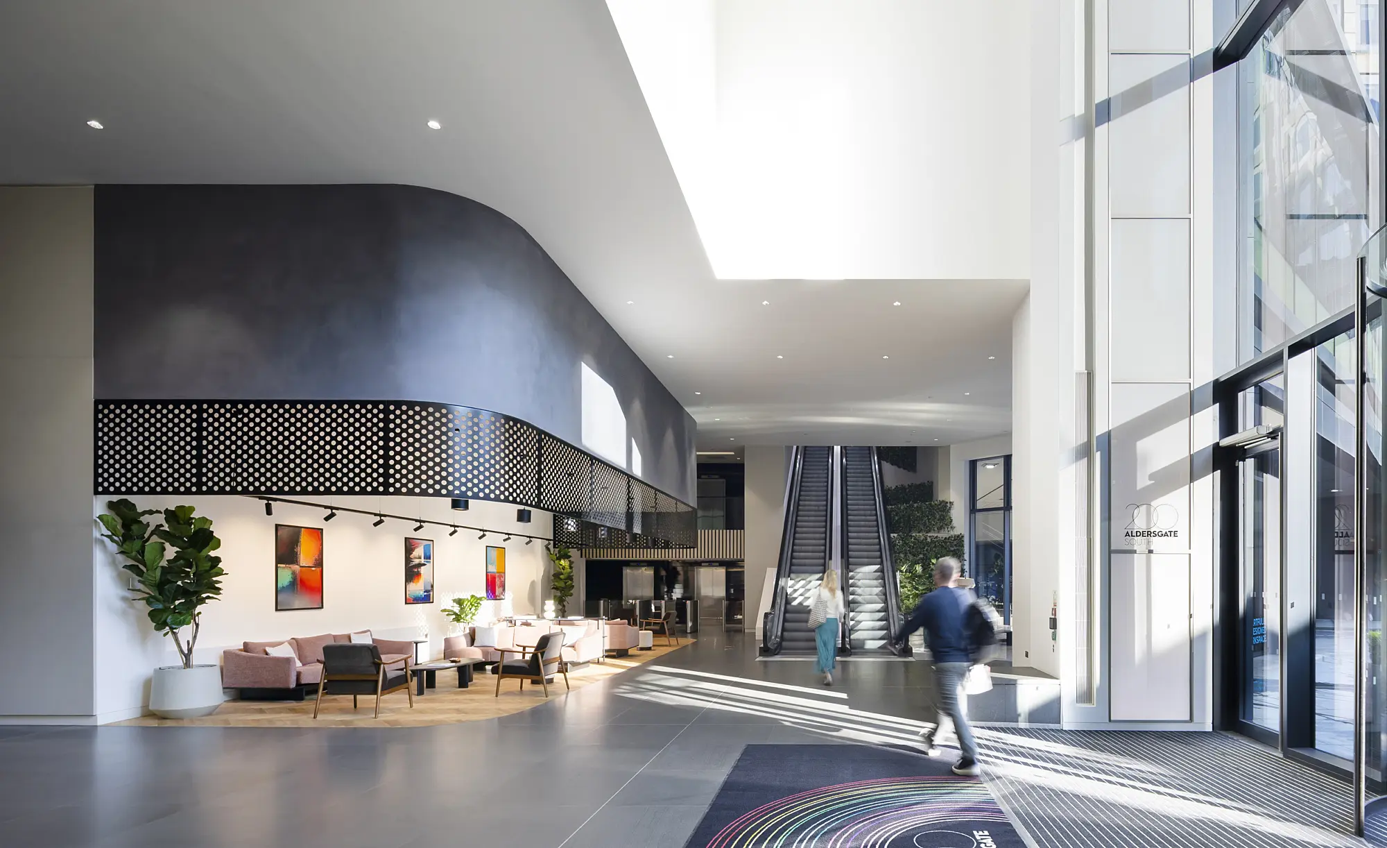 A bright, modern lobby with soaring ceilings and expansive windows that fill the space with natural light. Multiple seating areas feature chairs and sofas arranged for comfort and conversation. Vibrant artwork decorates the walls, while potted plants introduce touches of greenery. A tiled floor leads to a large entrance mat with a colorful circular design. In the background, an escalator ascends to an upper level, and a few people move through the space, adding a sense of activity.