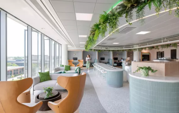 A bright, modern office designed with openness and comfort in mind. Large windows bring in natural light, illuminating tan leather chairs and grey sofas accented with green cushions. A long counter with bar stools offers a casual workspace, while a curved reception desk clad in light blue tiles adds a welcoming touch. Hanging greenery from the ceiling introduces natural elements into the space. Two people move through the background, contributing to the sense of activity.