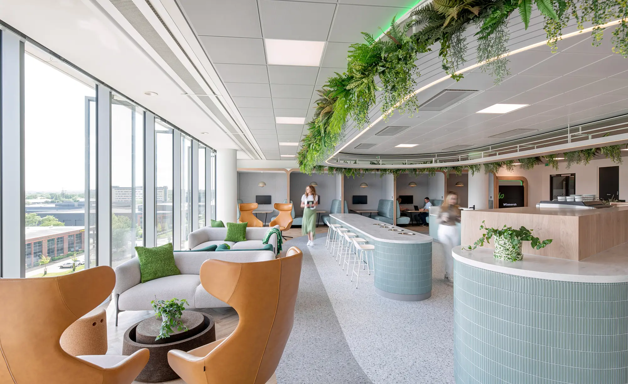 A bright, modern office designed with openness and comfort in mind. Large windows bring in natural light, illuminating tan leather chairs and grey sofas accented with green cushions. A long counter with bar stools offers a casual workspace, while a curved reception desk clad in light blue tiles adds a welcoming touch. Hanging greenery from the ceiling introduces natural elements into the space. Two people move through the background, contributing to the sense of activity.