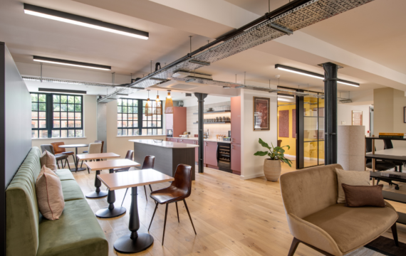Contemporary office breakout space with exposed steel beams, pink-toned kitchen cabinetry, soft seating, wooden flooring, and café-style tables beneath industrial lighting.
