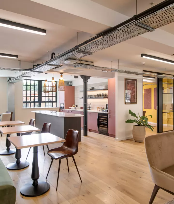 Contemporary office breakout space with exposed steel beams, pink-toned kitchen cabinetry, soft seating, wooden flooring, and café-style tables beneath industrial lighting.