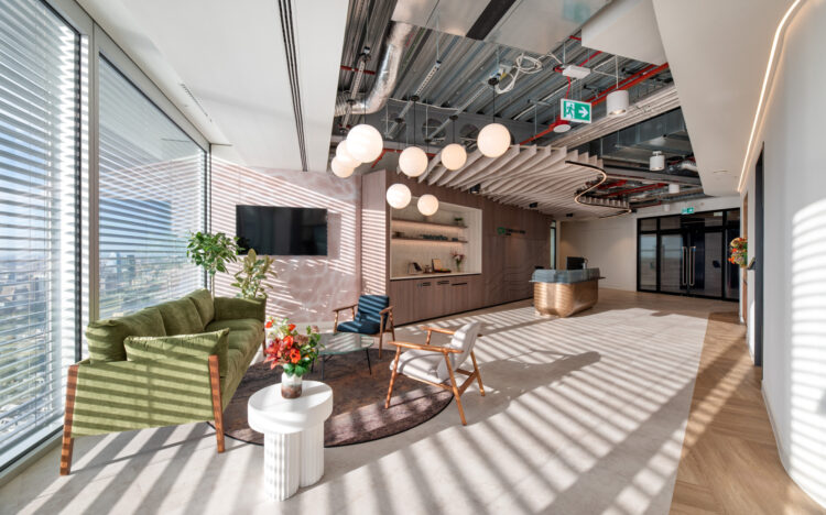 A modern office lobby area with large windows on the left side, partially covered by blinds, allowing natural light to filter in. The seating area includes a green sofa and two chairs arranged around a small round table, which holds a vase of flowers. The ceiling features exposed piping and several spherical pendant lights. In the background, a reception desk with a computer monitor is visible, along with shelving units behind it. The floor is finished with light-coloured tiles, contributing to the clean and welcoming atmosphere.