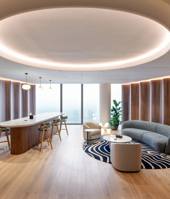 A stylish and modern office lounge area featuring a curved grey sofa that forms a semi-enclosed booth-like seating arrangement around a round coffee table placed on a patterned rug. The space includes a sleek oval-shaped bar table with high stools, positioned under a large circular ceiling light fixture. Wooden flooring and wall paneling add warmth to the environment, while a tall indoor plant in the corner and large windows offering natural light enhance the inviting atmosphere. The word "UniCredit" is displayed on the wall to the left, contributing to the branded interior design.