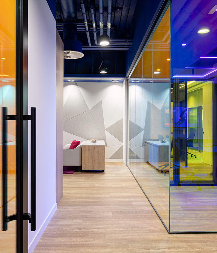 A modern office interior featuring glass walls and doors tinted in bold, vibrant colours including orange, yellow, blue, and purple. The space has a contemporary, open-plan layout with a light wood floor and an exposed ceiling showcasing industrial-style piping and lighting fixtures. In the background, people are walking through the corridor, adding a sense of activity. A small seating area with a sofa and table is visible through one of the coloured glass partitions, contributing to a welcoming and collaborative atmosphere.