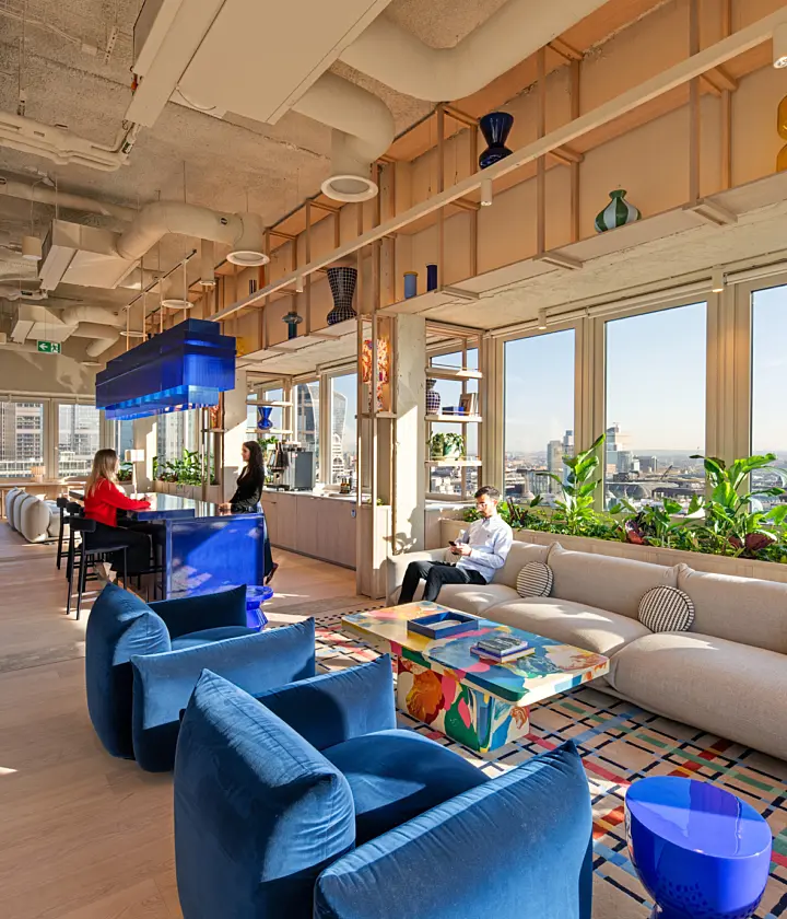 Modern office lounge with floor-to-ceiling windows overlooking the City of London skyline. The space features a large curved beige sofa, two blue lounge chairs, a colourful coffee table and abundant indoor plants. A glossy blue bar and matching suspended light fixture sit at the centre, while decorative glass vases line open shelving above. Three people occupy the space, with two seated at the bar and one seated on the sofa. Exposed ceilings, light wood finishes and natural daylight create a bright, contemporary workplace setting.