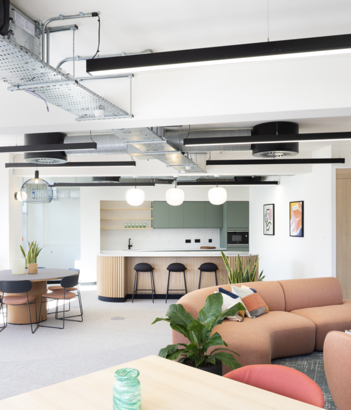A modern open-plan office featuring industrial-style exposed ceiling ducts and sleek black linear light fixtures. The space includes a kitchen area with light wood cabinetry, white countertops and black bar stools. A round dining table with four chairs sits near a large window that floods the room with natural light. The foreground showcases a peach-coloured L-shaped sofa surrounded by green plants and small tables, creating a relaxed lounge area. Framed artwork adds visual interest to the neutral-toned walls.