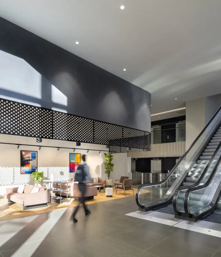 A modern indoor office space featuring a relaxed seating area with sofas, chairs and tables, accented by colourful wall art. An escalator on the right leads to an upper level, surrounded by greenery. Large windows fill the space with natural light, creating a bright, welcoming atmosphere.