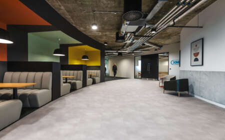 Modern office breakout area with an open industrial ceiling featuring exposed ductwork and lighting. On the left, a row of upholstered booth seating in neutral tones lines the wall, each booth paired with a small round wooden table and set within colourful alcoves in shades of green, yellow and orange. Pendant lights hang above the seating. The centre of the space has a wide, light grey floor leading towards a corridor, where one person is walking in the distance. On the right, there is a lounge area with a cushioned chair, a small table and framed abstract artwork on a white and grey wall.