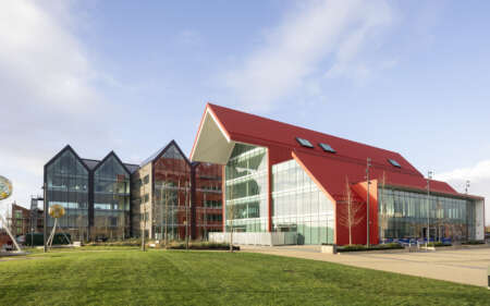 Modern campus-style building with a red pitched roof and large glass façades, connected to a darker grey, multi-gabled glass structure on the left. The buildings sit beside a wide paved walkway and a neatly maintained green lawn in the foreground. Sculptural metal frames holding spherical artworks stand on the grass. The scene is brightly lit by sunlight under a clear blue sky with light clouds, highlighting the clean lines, reflective windows and contemporary architectural design.