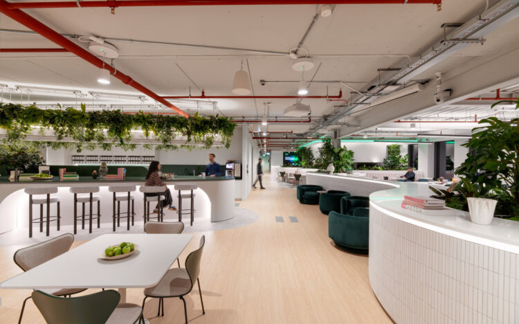 A spacious modern workplace with light wood flooring, white curved counters and abundant greenery. A central bar-style area with stools sits beneath a ceiling feature covered in hanging plants. People are seated at tables and lounge chairs throughout the open space. Overhead pipes and lighting are exposed, and the setting has a bright, contemporary feel.