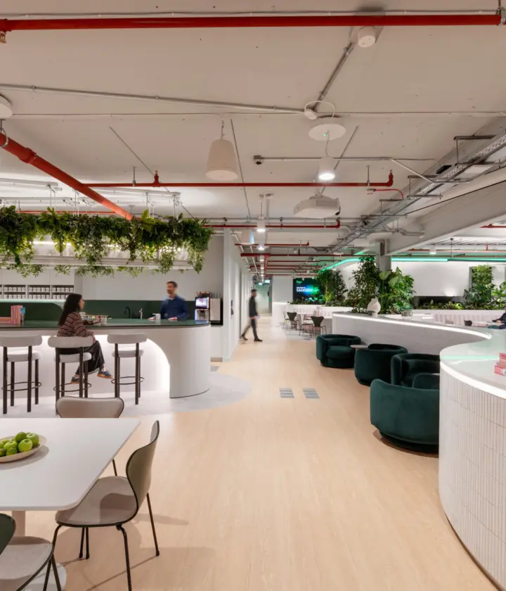 A spacious modern workplace with light wood flooring, white curved counters and abundant greenery. A central bar-style area with stools sits beneath a ceiling feature covered in hanging plants. People are seated at tables and lounge chairs throughout the open space. Overhead pipes and lighting are exposed, and the setting has a bright, contemporary feel.