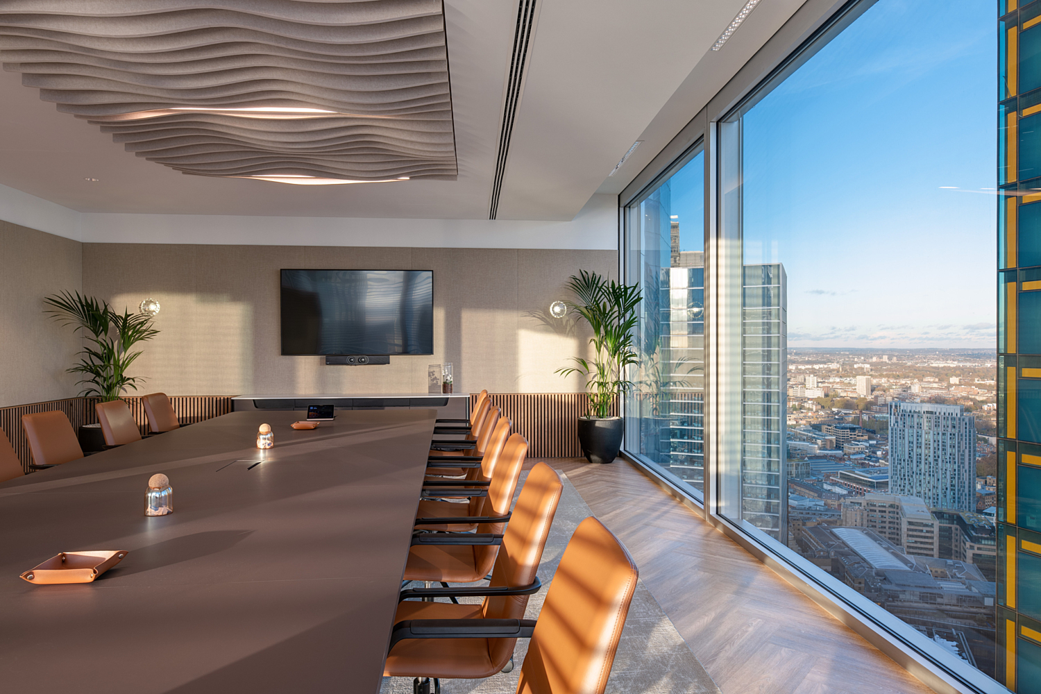 A modern conference room featuring a long table surrounded by multiple chairs. Large windows along one side provide a view of London and allow natural light to enter the space. The room includes a wall-mounted flat-screen television, potted plants placed in the corners, and distinctive ceiling lights with a wavy design, contributing to the contemporary aesthetic.