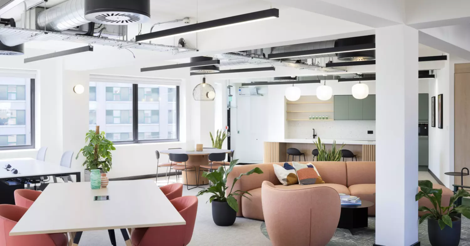 An open-concept office space designed for flexibility and comfort. The room features a mix of seating options, including tables with chairs and a pink sofa set. Green plants are placed throughout, adding freshness to the environment. Exposed ductwork and modern lighting fixtures give the ceiling an industrial edge. Large windows bring in natural light, and a kitchenette with green cabinets and hanging lights sits in the background, offering convenience and style.