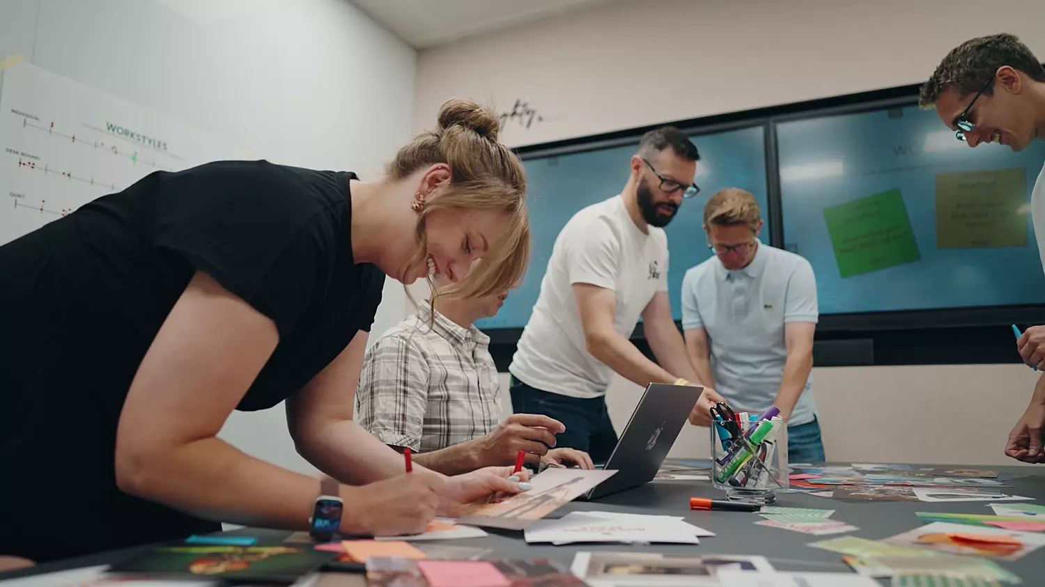 A dynamic team environment captured during a brainstorming planning session. A group of people are gathered around a table scattered with papers, photographs and stationery. In the background, a whiteboard displays text and diagrams, reinforcing the collaborative nature of the activity. The scene reflects a hands-on, creative process in progress, highlighting teamwork and shared focus.
