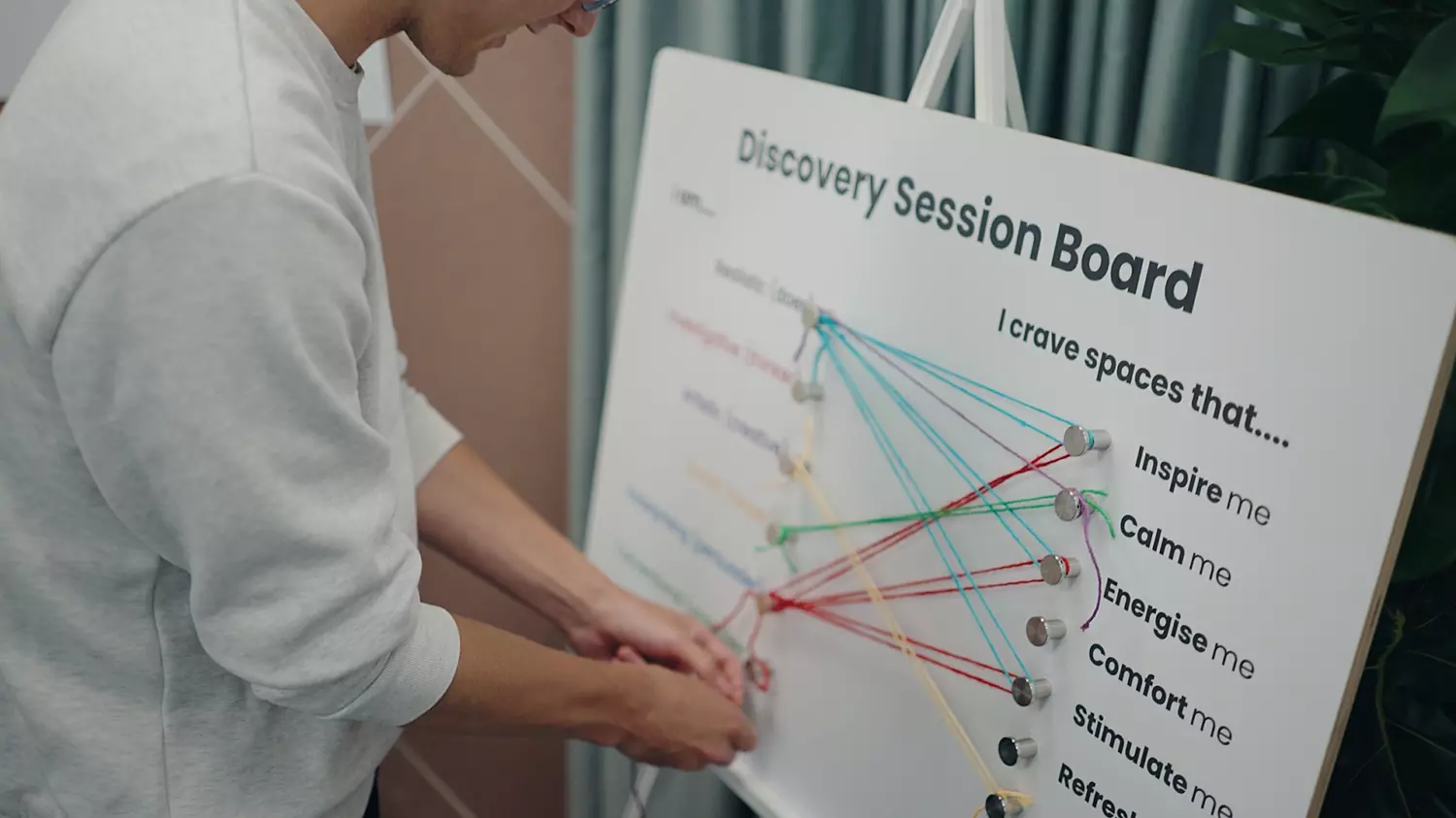 A focused moment captured during a discovery session. A person engages with a board titled Discovery Session Board, which features colored strings linking various traits and preferences. The board explores personal attributes such as being realistic, imaginative, analytical and reflective, alongside spatial needs like inspiration, calm, energy, comfort and stimulation. The visual layout suggests a structured yet creative approach to understanding individual working styles and environmental preferences.