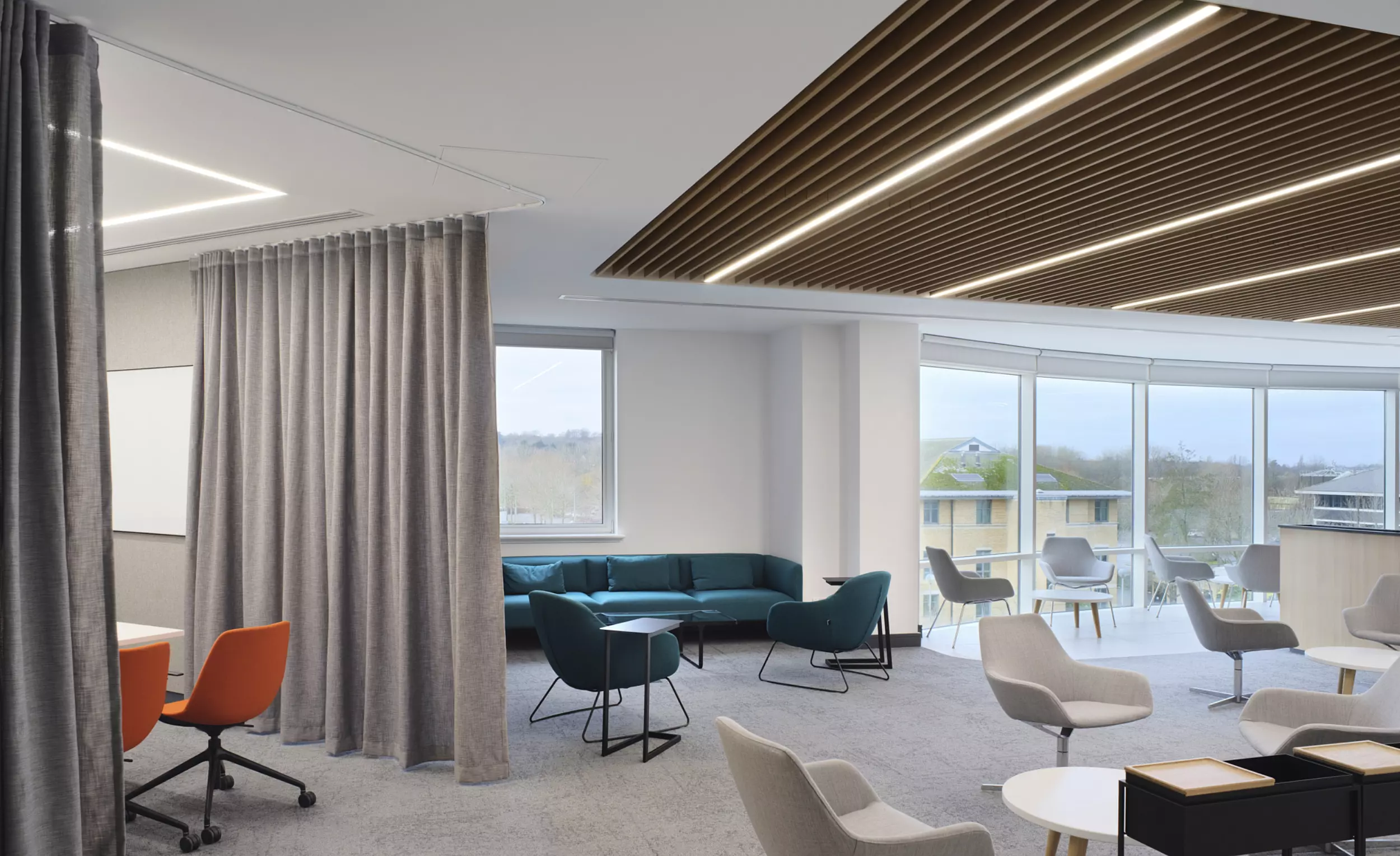 An office space arranged with a variety of seating options, including orange swivel chairs, white and grey lounge chairs, and teal sofas, supporting flexible use and informal collaboration. A curtain partition provides a soft spatial divide, allowing for adaptable zoning within the open-plan layout. A large window introduces natural light, enhancing the usability of the space. The ceiling features wooden slats with integrated lighting, and a whiteboard is positioned at the rear, indicating a designated area for brainstorming or presentations.