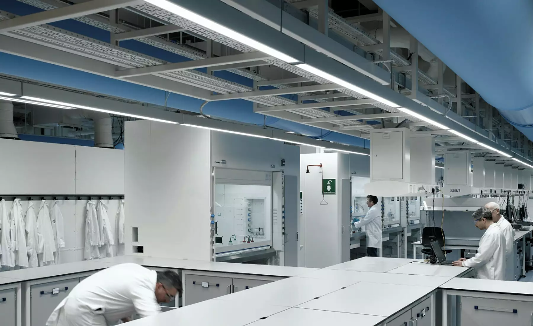 A clean, well-organised laboratory with multiple workstations and individuals in white lab coats engaged in various tasks. The space features workbenches, storage cabinets, and drawers arranged for efficiency. Lab coats hang neatly on hooks along the left wall. The ceiling reveals exposed piping and ventilation ducts, contributing to an industrial aesthetic that supports functionality and airflow.