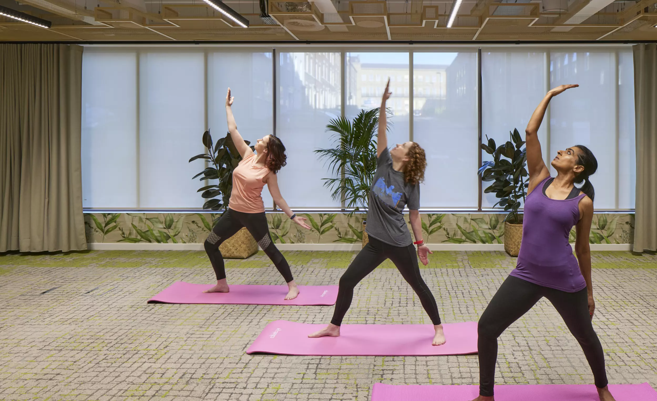 Three people are taking part in a yoga class in the office. They are standing on pink yoga mats, holding a side-reaching pose with one arm extended upwards and the other resting behind them. The room has large windows with blinds, potted plants, green and beige carpet tiles and a ceiling with modern lighting and acoustic panels.