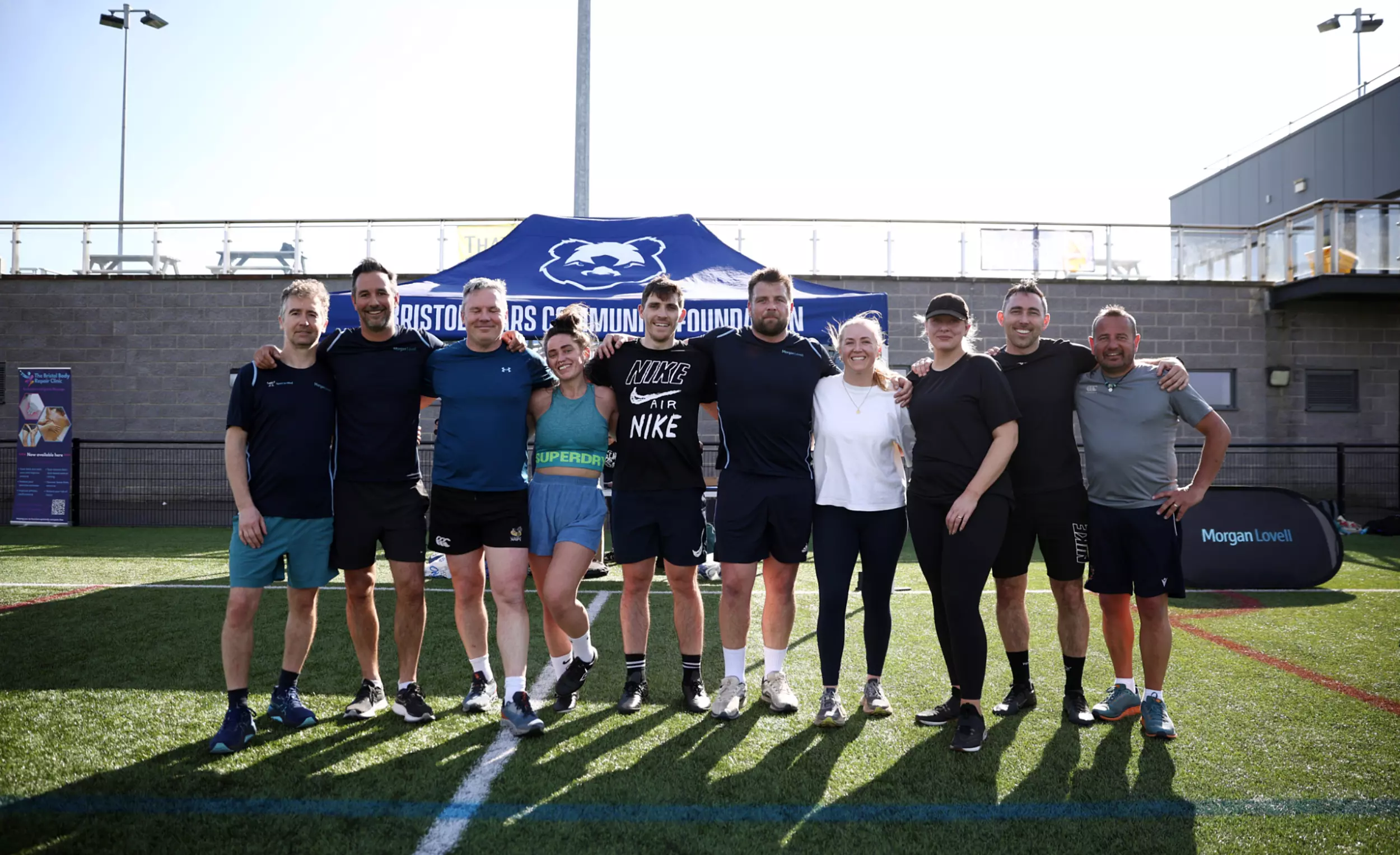 A group of ten people stand side by side on an outdoor sports field, smiling for the camera. They are dressed in sportswear and have just taken part in a charity rugby event. Behind them is a blue tent with the logo and text "Bristol Bears Community Foundation" and a banner on the right with the logo "Morgan Lovell." The setting is sunny, with a clear sky and a modern building with a balcony in the background.
