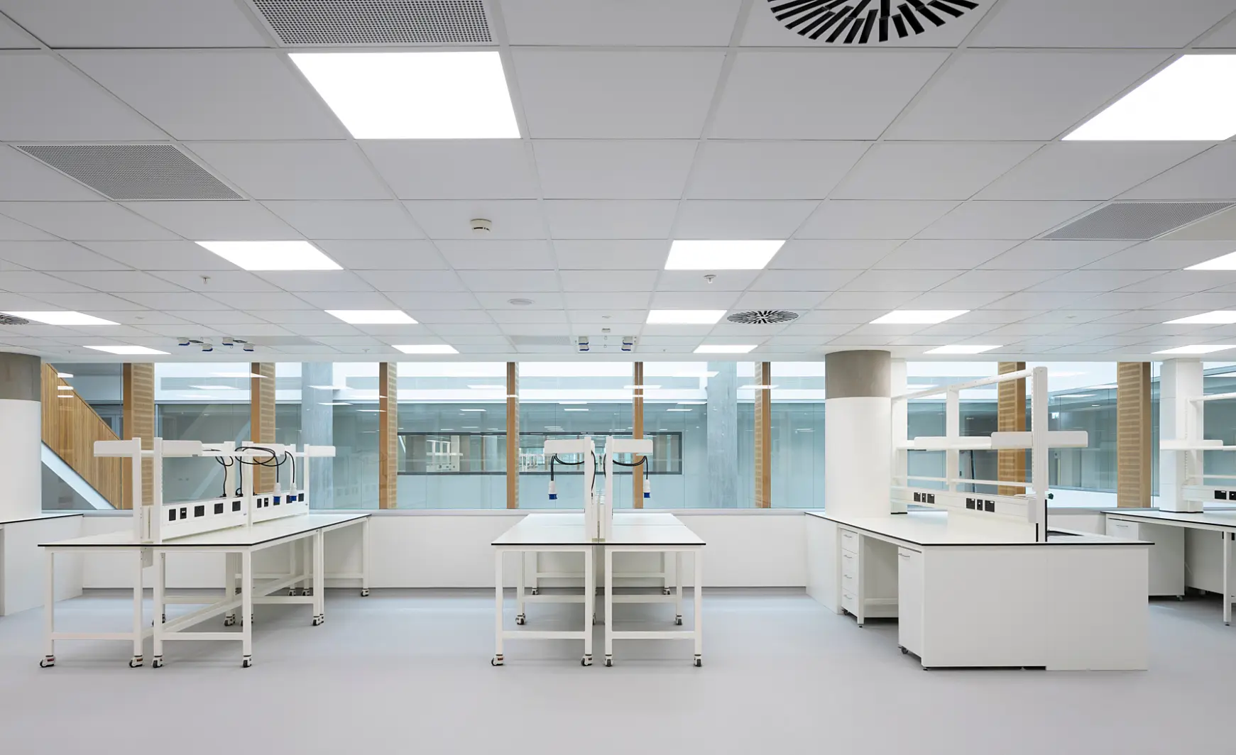 A bright, clean laboratory featuring multiple workstations with white countertops and matching cabinetry. Large windows at the back of the room allow natural light to fill the space. The ceiling is fitted with evenly spaced lighting panels and visible ventilation systems. The overall layout is minimalistic, with a focus on functionality and efficient use of space.