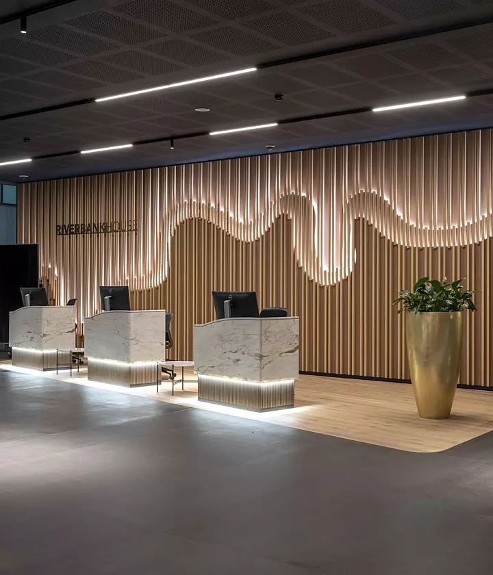 A modern, stylish interior space featuring contemporary furniture including blue armchairs and sofas. A patterned rug and a large potted plant are in the foreground. The background includes wooden wall panelling, ceiling lights and a partition with vertical slats dividing the room into separate areas.