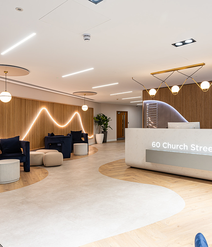 A modern office reception area with a clean, minimalist design. The space features a light wood and white colour scheme with a wave-like LED light pattern on a slatted wooden wall. The reception desk is white with "60 Church Street" lit up on the front. There are navy blue armchairs, round pouffes and small white tables arranged in seating areas. Large potted plants add greenery and contemporary pendant lights hang from the ceiling. The flooring blends light wood and white finishes in a flowing pattern.