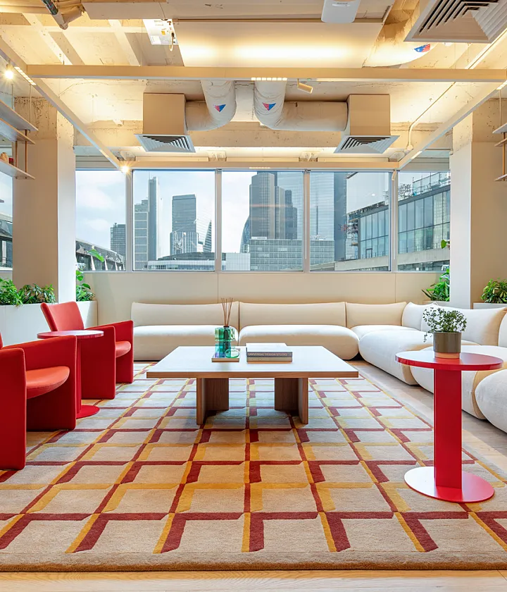 A bright office lounge area with large windows offering views of city buildings. The space includes a long white L-shaped sectional sofa arranged around a central coffee table, flanked by two red chairs and two small round tables, one red and one white. A patterned rug in beige and red covers the floor. Green plants line the edge of the seating area, and the ceiling features exposed ductwork and lighting fixtures.