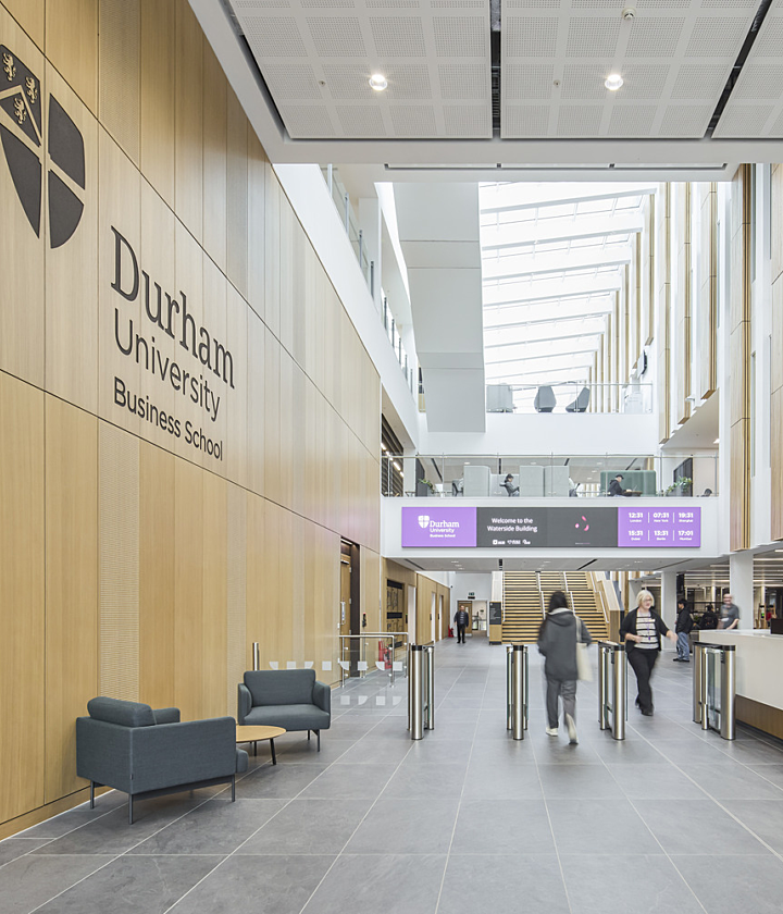 A contemporary interior space within Durham University Business School, featuring wooden panelled walls and a high ceiling with integrated lighting. Large windows allow ample natural light to flood the area. The space includes modern seating arrangements with chairs and sofas, an information desk on the right, and turnstiles near the entrance. The university’s logo and name are displayed prominently on the left wall.