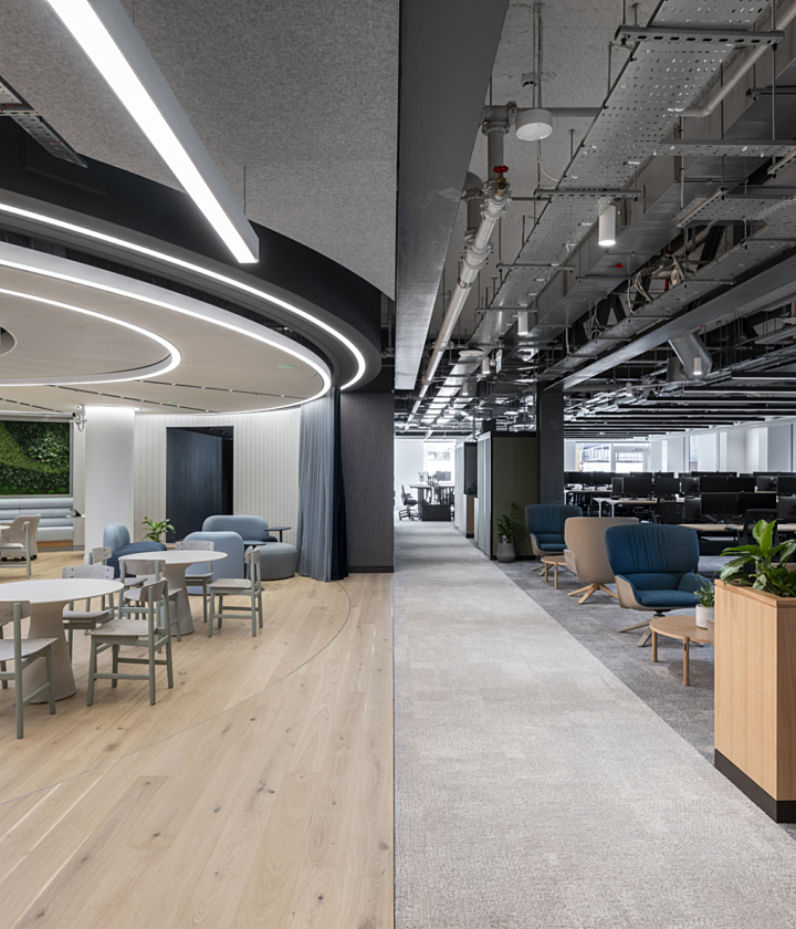 A modern office interior split into two distinct areas. On the left side, there's a bright, stylish lounge space with light wood flooring, soft pastel chairs, round tables, and a green living wall in the background. The ceiling features circular recessed lighting that adds a sleek, futuristic feel. On the right side, there's an open-plan workspace with grey carpet, rows of black computer monitors on desks, and exposed industrial ceiling elements like pipes and cables. This area also includes some casual seating with blue and beige armchairs, coffee tables, and planter boxes with greenery.