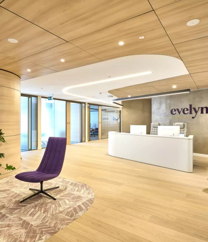 Modern office reception with a minimalist design featuring light wood flooring and curved timber wall panels. The ceiling has matching timber finishes with recessed lighting and integrated acoustic panels. A sleek white reception desk sits against a textured wall with the Evelyn Partners logo. The space includes contemporary furniture such as a purple lounge chair, a patterned rug, and indoor plants for a natural touch. Glass partitions in the background lead to meeting rooms, allowing natural light to flow through