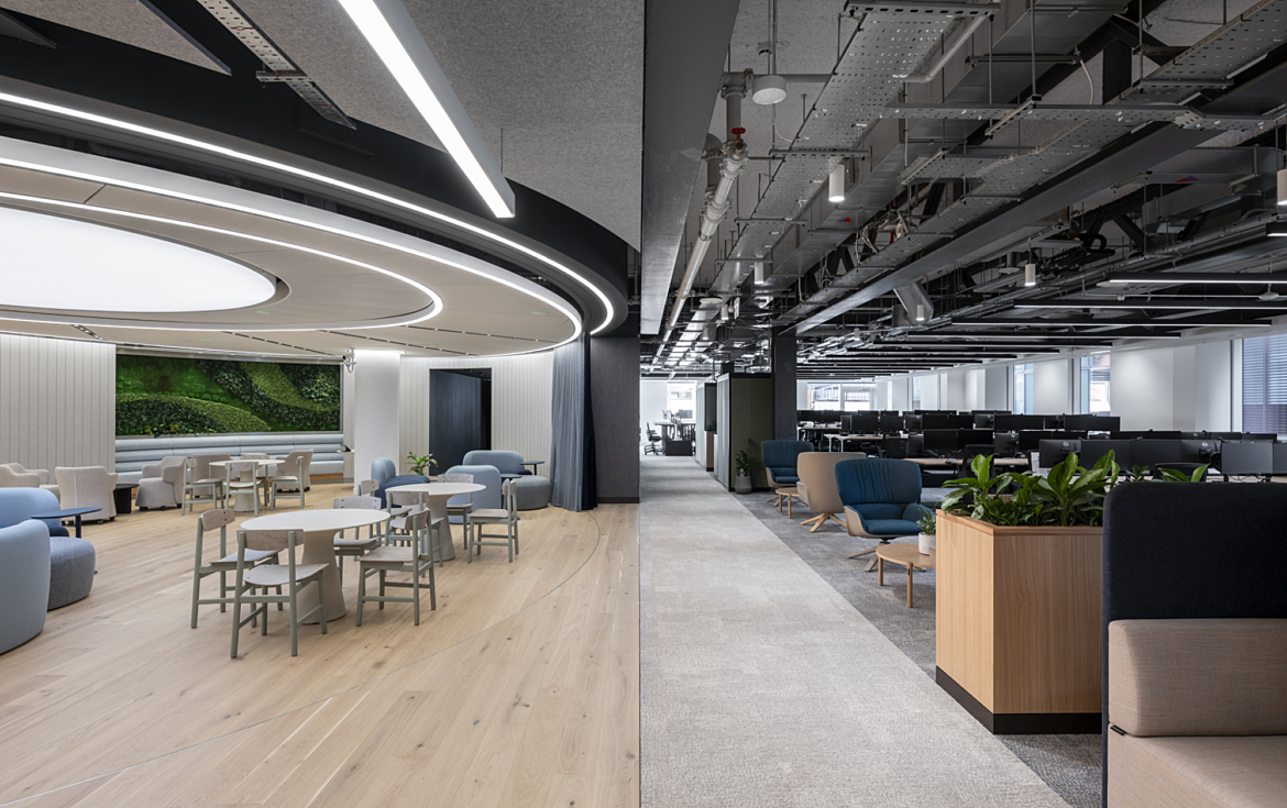 A modern office interior split into two distinct areas. On the left side, there's a bright, stylish lounge space with light wood flooring, soft pastel chairs, round tables, and a green living wall in the background. The ceiling features circular recessed lighting that adds a sleek, futuristic feel. On the right side, there's an open-plan workspace with grey carpet, rows of black computer monitors on desks, and exposed industrial ceiling elements like pipes and cables. This area also includes some casual seating with blue and beige armchairs, coffee tables, and planter boxes with greenery.