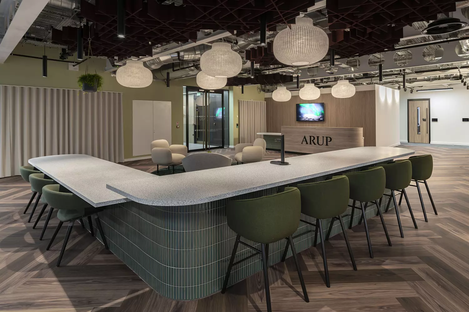 A contemporary office interior featuring a large, curved communal table with a sleek white surface, surrounded by green upholstered chairs. Overhead, spherical pendant lights hang from a ceiling with exposed industrial-style piping and ductwork. The floor is laid with herringbone-patterned wooden flooring. In the background, a glass-walled meeting room and a reception area are visible, with the word "ARUP" displayed on the front desk. The walls are painted in neutral tones, with vertical wooden slats adding texture and warmth to one section.