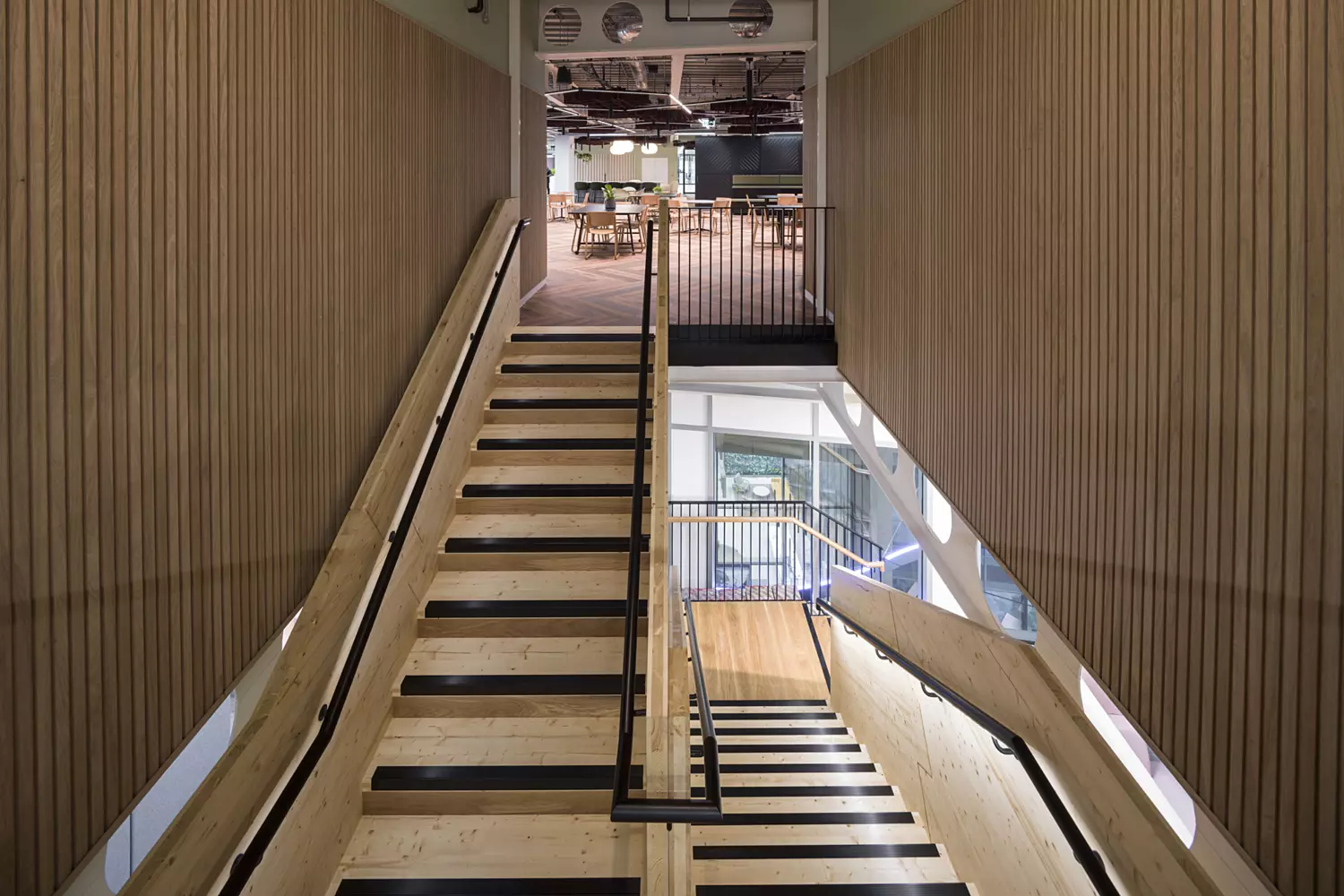 A contemporary indoor staircase with wooden treads and sleek black railings, splitting into two directions at the base. The surrounding walls are clad in vertical wooden slats, adding warmth and texture. At the top of the stairs, a glass partition reveals an open-plan workspace or lounge area furnished with tables and chairs.
