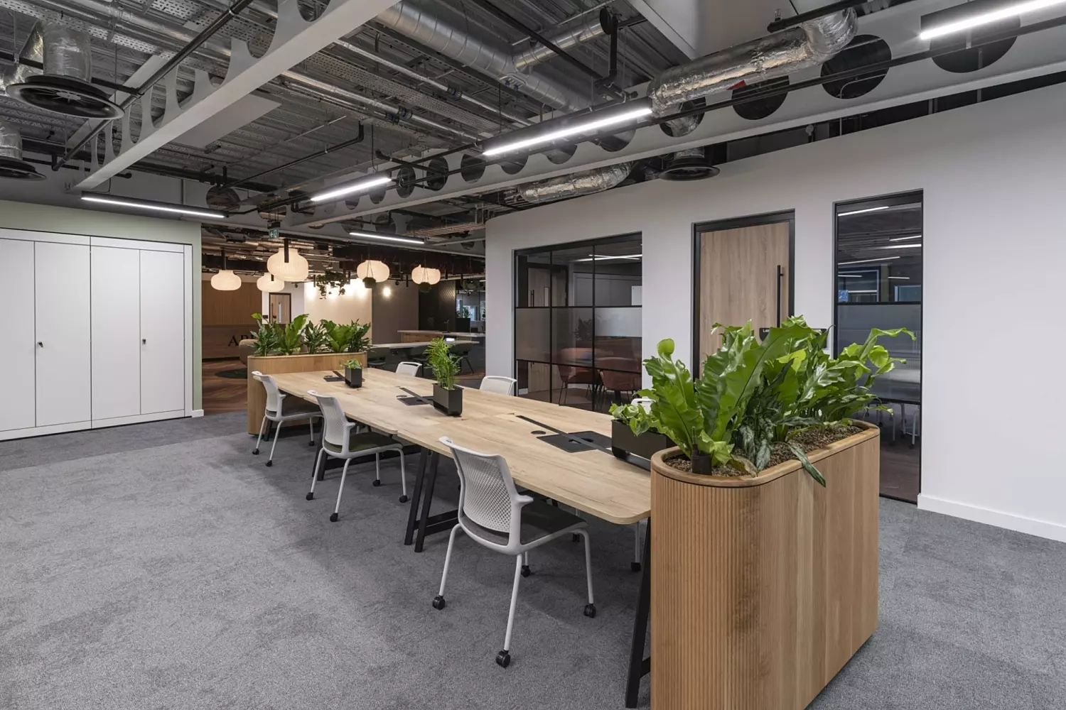 A modern open-plan office featuring a long wooden table at the centre, surrounded by chairs and adorned with potted plants both on the tabletop and at either end. The ceiling showcases exposed piping and industrial-style lighting fixtures. To the left, white storage cabinets line the wall, while to the right, a glass-walled meeting room or private office adds transparency and structure. The floor is finished with grey carpeting, contributing to the space’s clean and professional aesthetic.
