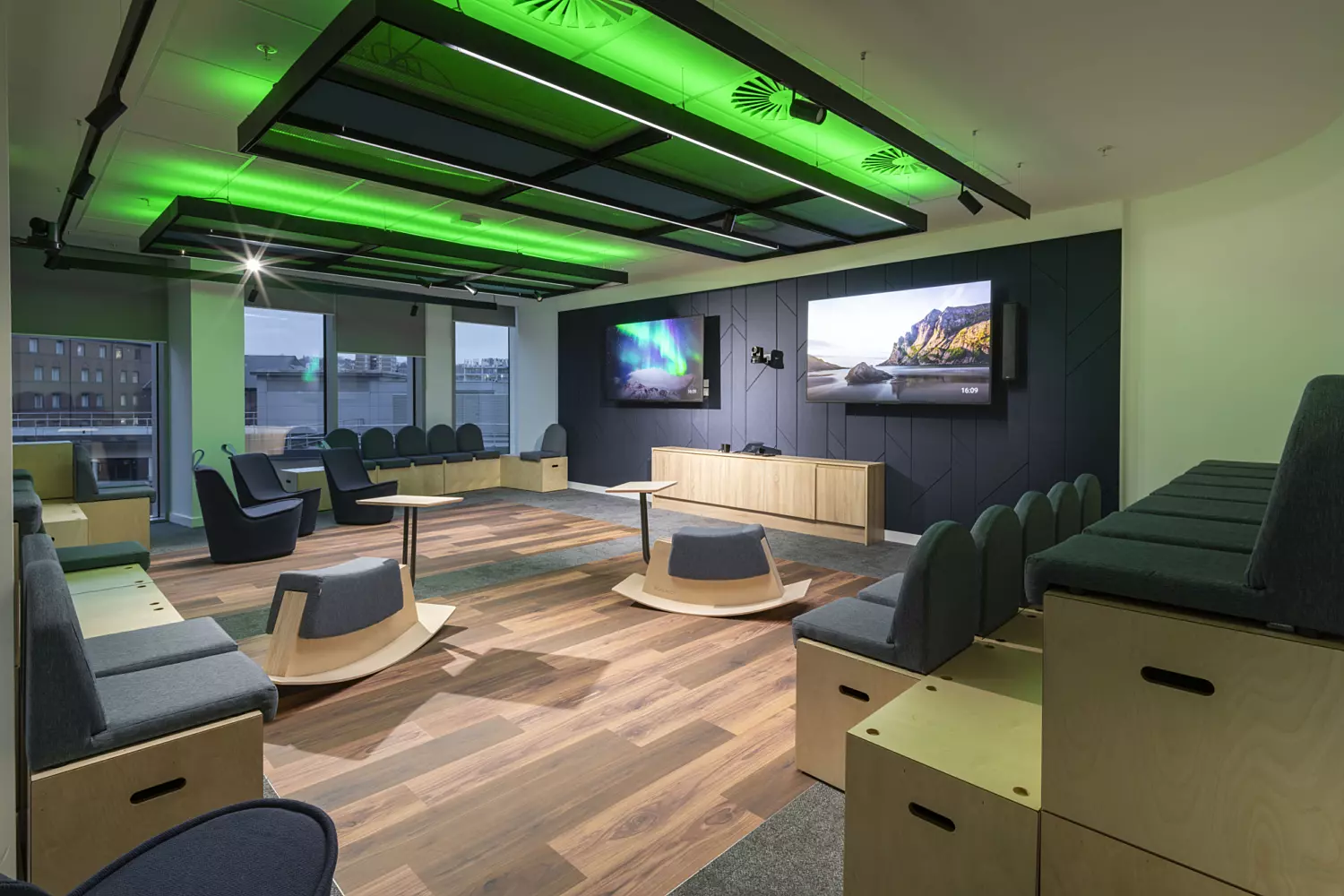 A stylish, modern room with wooden flooring and contemporary furnishings. Chairs are arranged in a semi-circle, with additional seating lining the walls. Two large flat-screen televisions are mounted on the wall, displaying scenic visuals. The ceiling is fitted with green lighting panels, adding a vibrant and dynamic accent. Natural light enters through windows, enhancing the room’s bright and welcoming atmosphere.