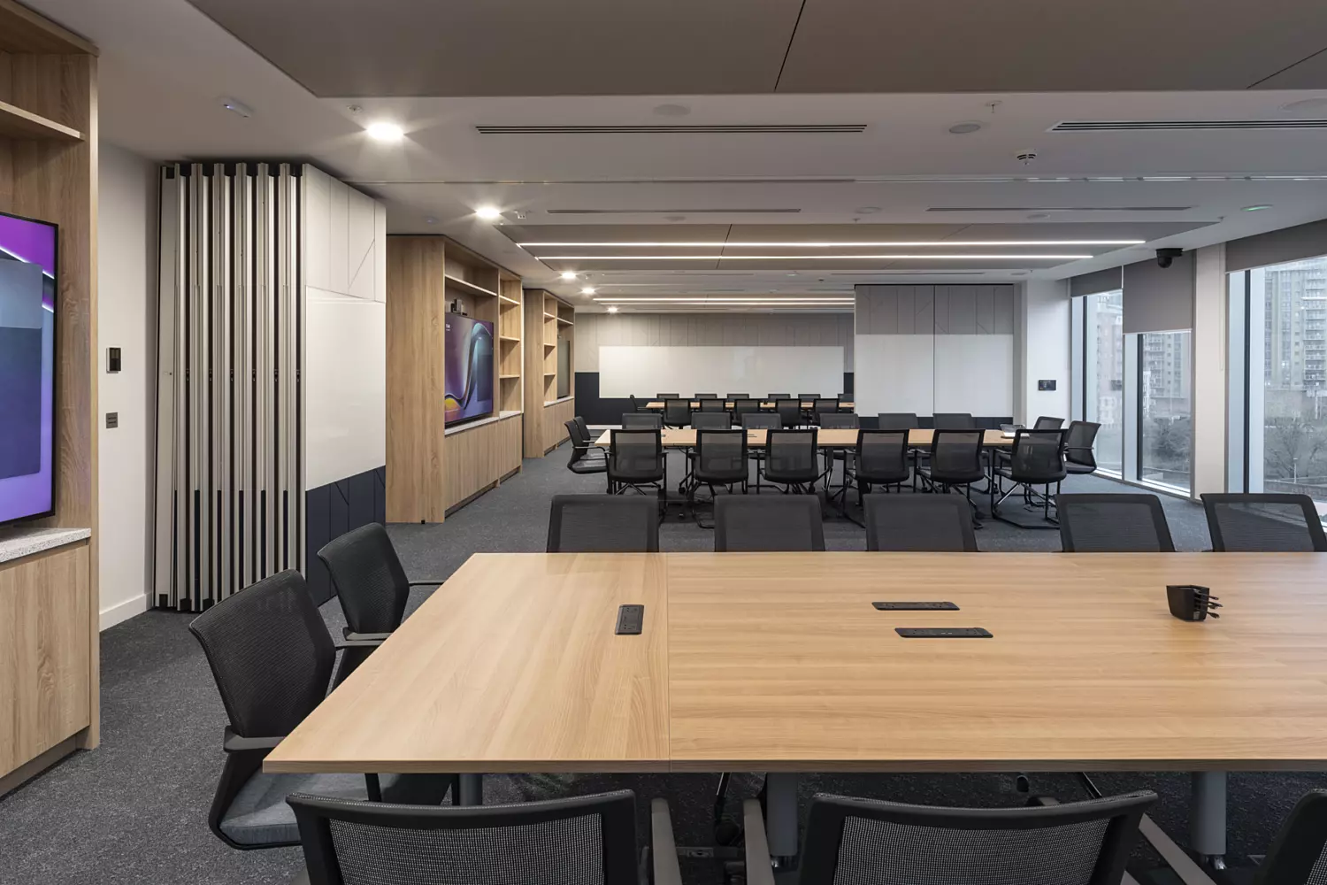 A modern conference room featuring a large wooden table in the foreground, surrounded by black mesh chairs. Behind it, rows of similar chairs are arranged in a classroom-style layout, facing the front. The space is well-lit with natural light from large windows along one side, complemented by recessed ceiling lights and acoustic panels. Two large display screens are mounted on opposite walls, enhancing the room’s functionality for presentations and meetings.