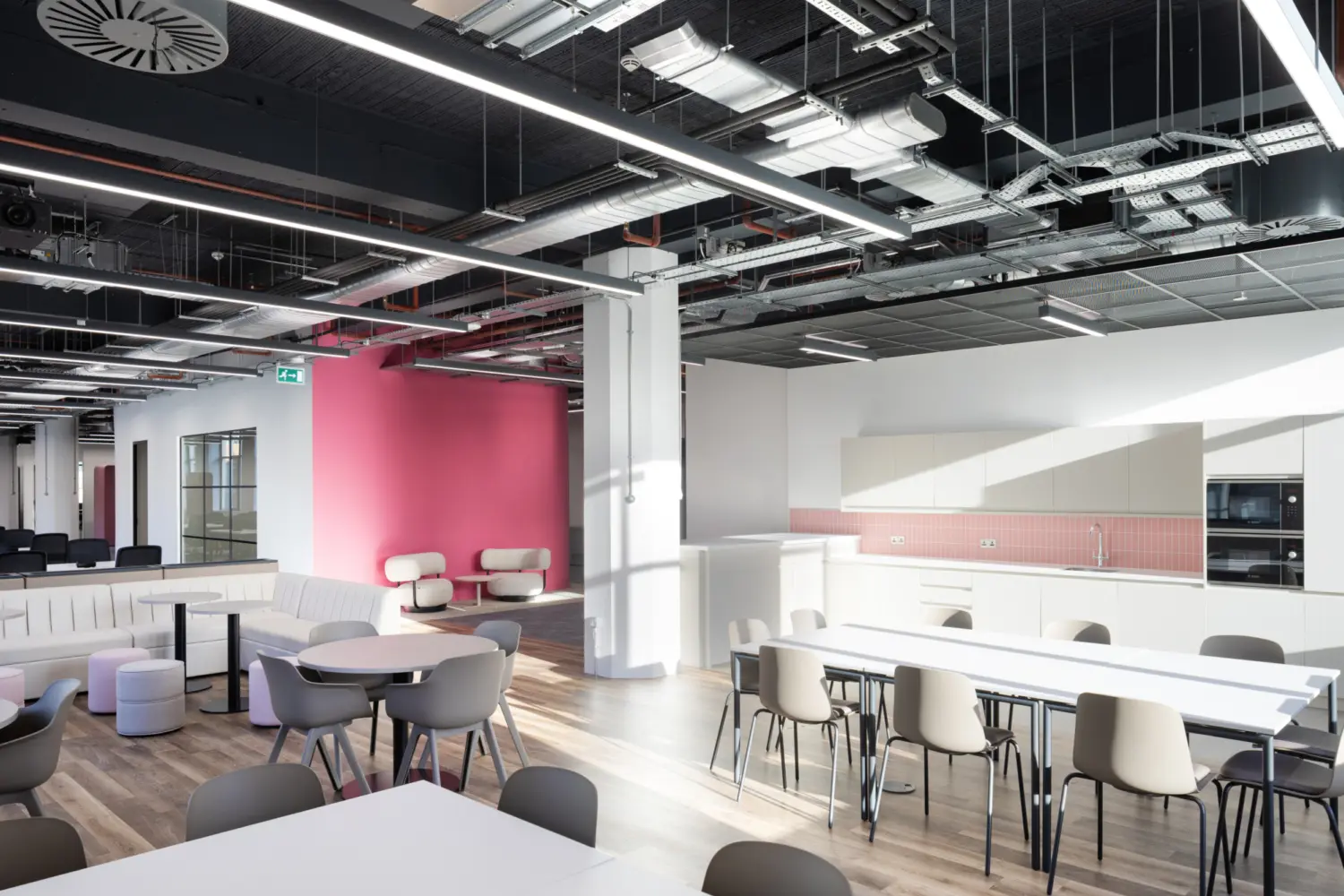 Modern open plan office breakout and kitchen area with a clean, contemporary fit out. The space features long white dining tables with light grey chairs in the foreground, alongside smaller round tables and soft seating to the left. At the back right, a white kitchenette includes integrated appliances, cabinets and a pink tiled splashback. A bold pink feature wall sits behind two white lounge chairs, adding contrast to the otherwise neutral palette. The ceiling is exposed, showing ventilation, cable trays and linear LED lighting, while large windows and overhead lights create a bright, evenly lit environment. The flooring is a mix of wood effect vinyl and darker carpet in the seating area.
