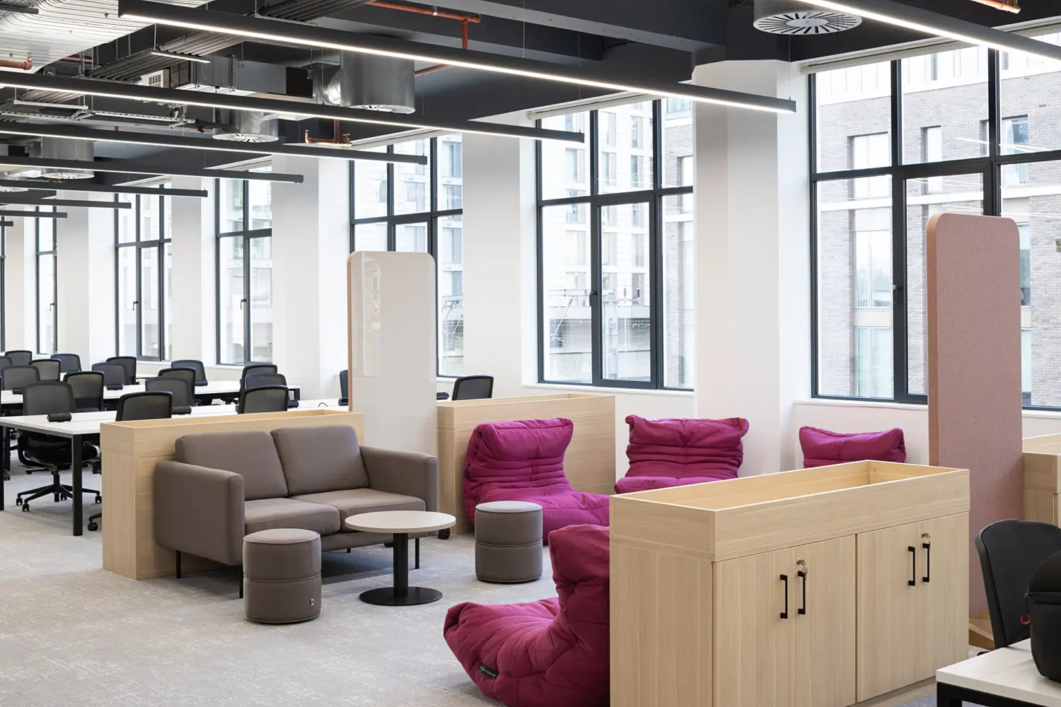Modern open plan office breakout and workspace area with a contemporary fit out and exposed ceiling showing ductwork, cable trays and linear LED lighting. Large floor‑to‑ceiling windows line the back wall, bringing in natural light and views of neighbouring buildings.  In the foreground, a soft seating area features a light grey sofa, two round ottomans and a small circular table, positioned beside a cluster of bright pink lounge chairs with a relaxed, low profile design. Light wood storage units and integrated planters help define the seating zone and create separation from surrounding areas.  To the left, rows of white desks with black task chairs form a structured workspace, arranged in lines across the open floor. The flooring is a light grey finish throughout, keeping the palette neutral and cohesive, while the pink seating introduces a bold accent.  The space combines collaborative workstations with informal seating, offering a mix of focused work areas and relaxed breakout zones within a bright, clean and modern office environment.