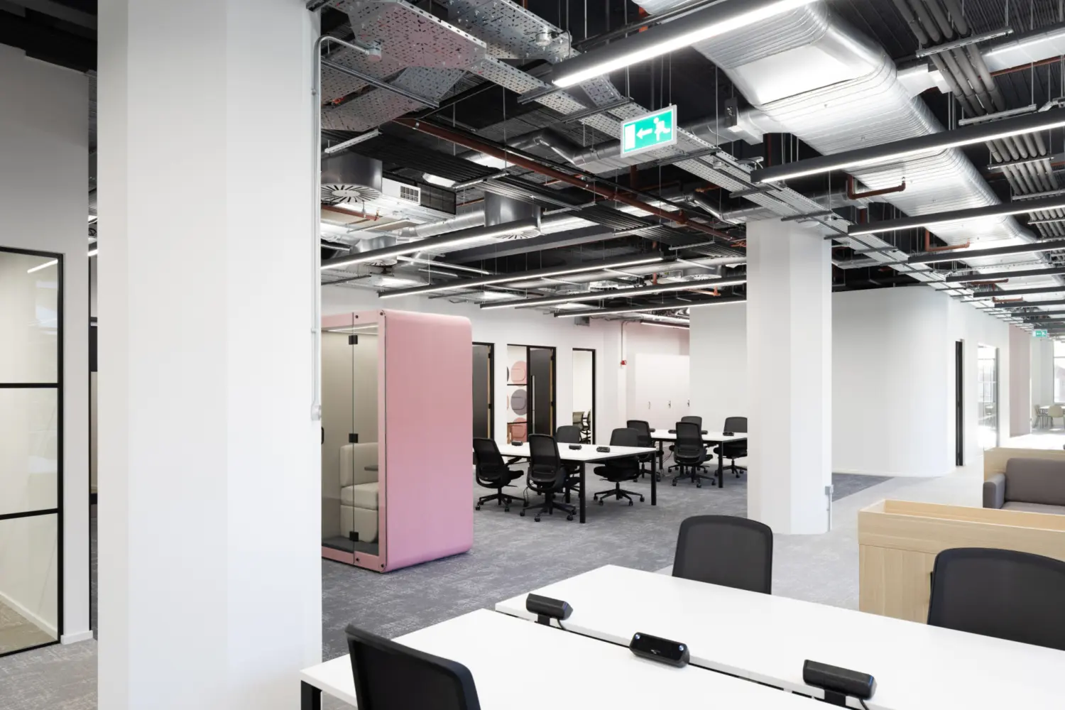 Modern open plan office workspace with a contemporary fit out featuring an exposed ceiling with visible ductwork, cable trays and linear LED lighting throughout. In the foreground, white bench desks are arranged with black task chairs and integrated desk power units. Large white structural columns run through the space, creating a sense of depth and division.  In the midground, a cluster of desks with black office chairs sits on grey carpet tiles, forming a collaborative working area. To the left, a glass meeting room with black-framed partitions is partially visible. Near the centre, a tall freestanding acoustic booth in soft pink adds colour and provides a private space within the open layout.  To the right, a transition to a breakout area is suggested by a light wood partition and soft seating. The overall colour palette is neutral with white, grey and black tones, accented by the pink booth. Bright, even lighting and clean lines give the space a functional and contemporary feel.